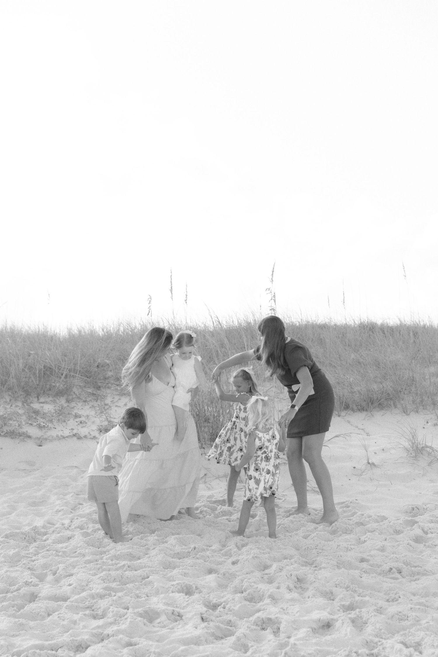 Black and white photo of a woman in a long dress with children playing on sandy beach dunes.