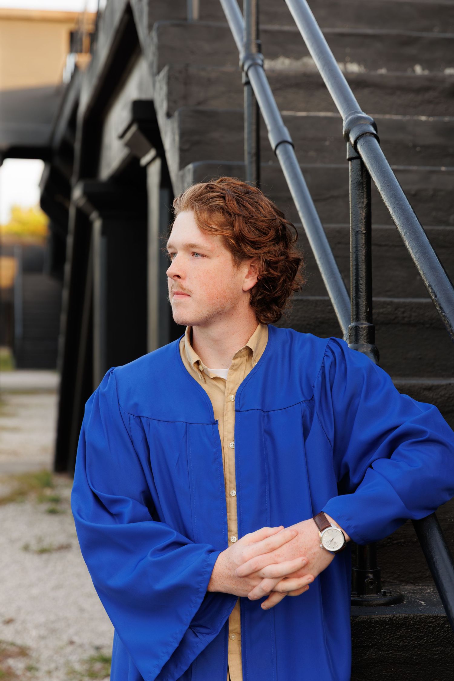 Young man in blue graduation gown poses outdoors near dark metal stairs, wearing a watch and looking to the side.