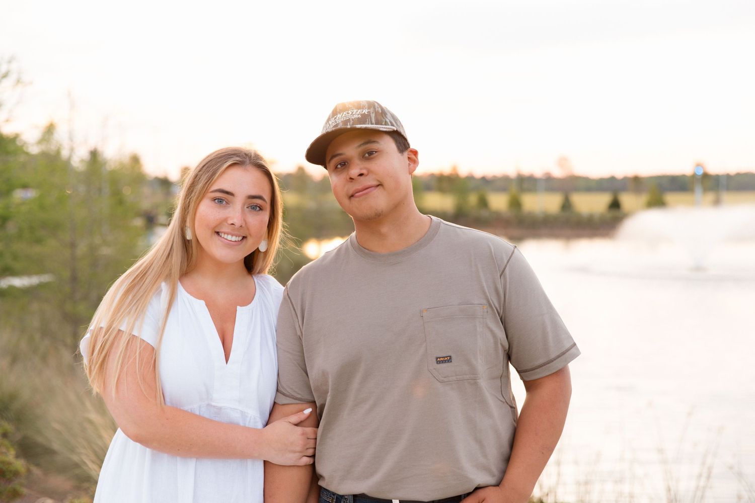 A young couple smiles together outdoors near a lake at golden hour, woman in white dress, man in gray t-shirt and camo cap.