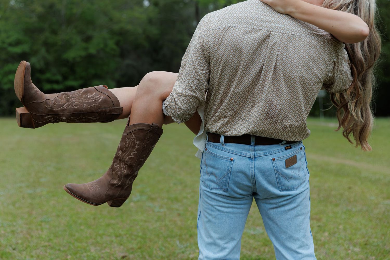 Man in light blue jeans carrying a woman wearing brown cowboy boots in a grassy outdoor setting.