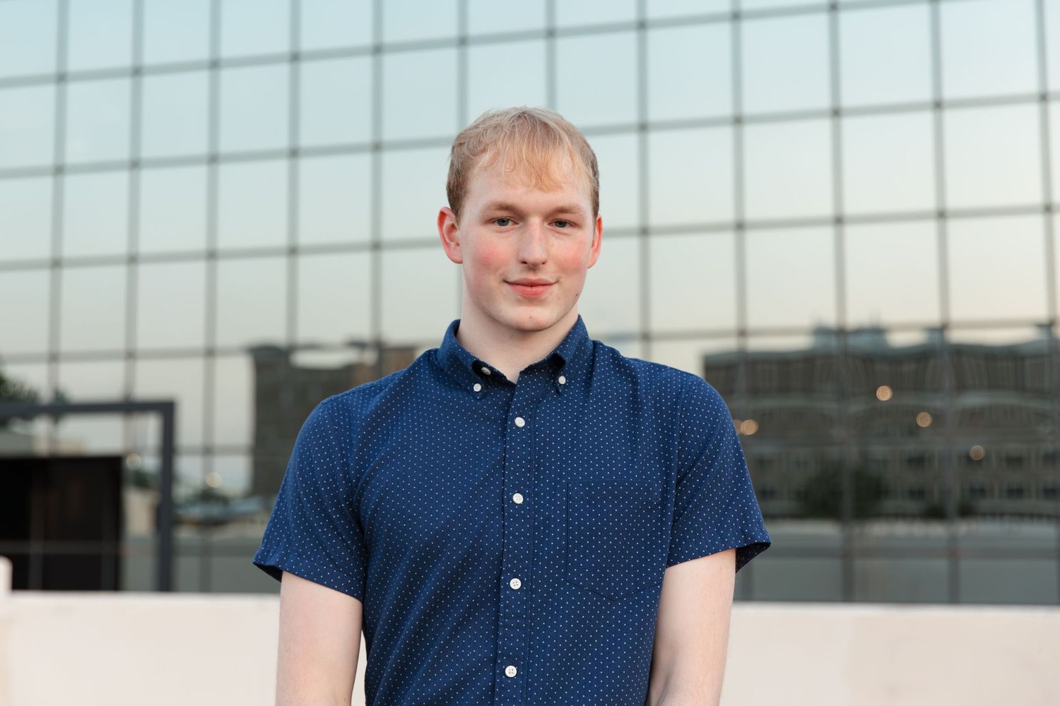 Young man in navy blue polo shirt smiling outdoors in front of modern glass building facade.