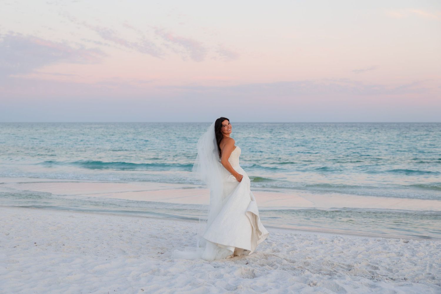 Bride in white gown and veil stands on sandy beach at sunset with turquoise ocean waves in background.