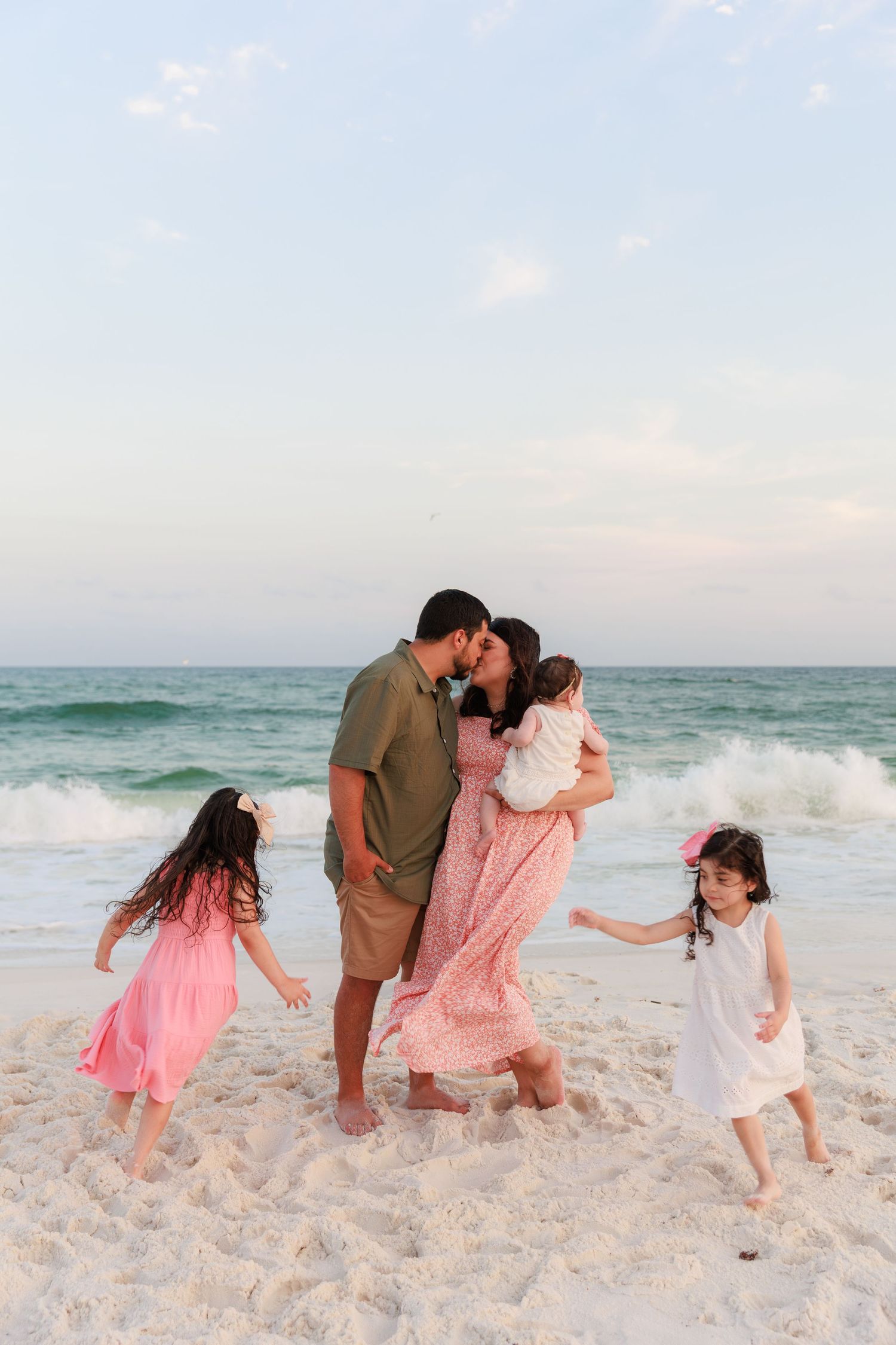 Parents share a kiss while holding baby as two daughters play around them on a sandy beach.