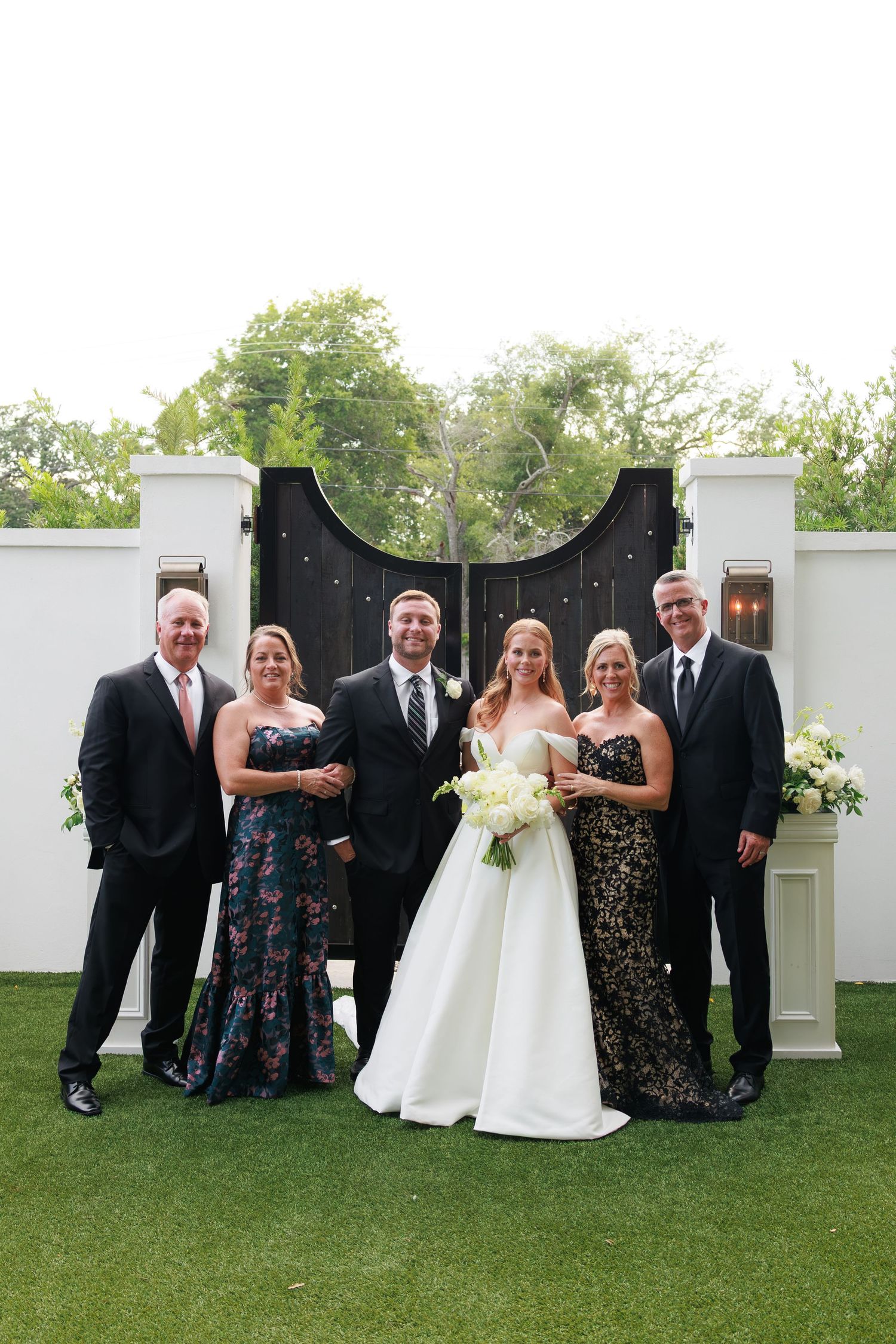 Bride and groom pose with family members in formal attire before elegant black gates at outdoor wedding venue.