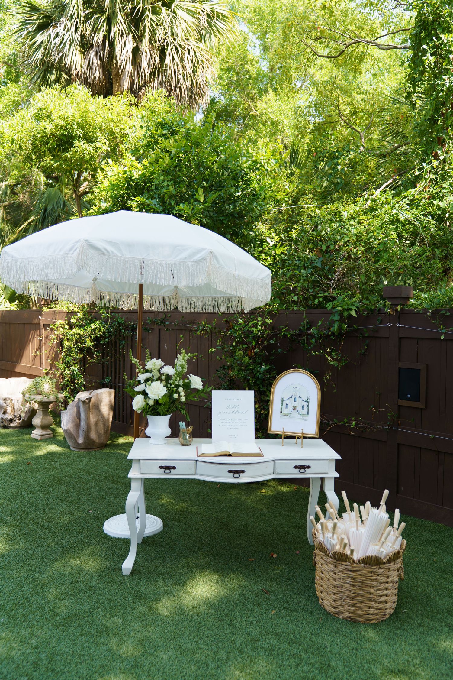 Elegant outdoor wedding welcome table with white umbrella, floral arrangement, guest book, and wicker basket on green lawn.