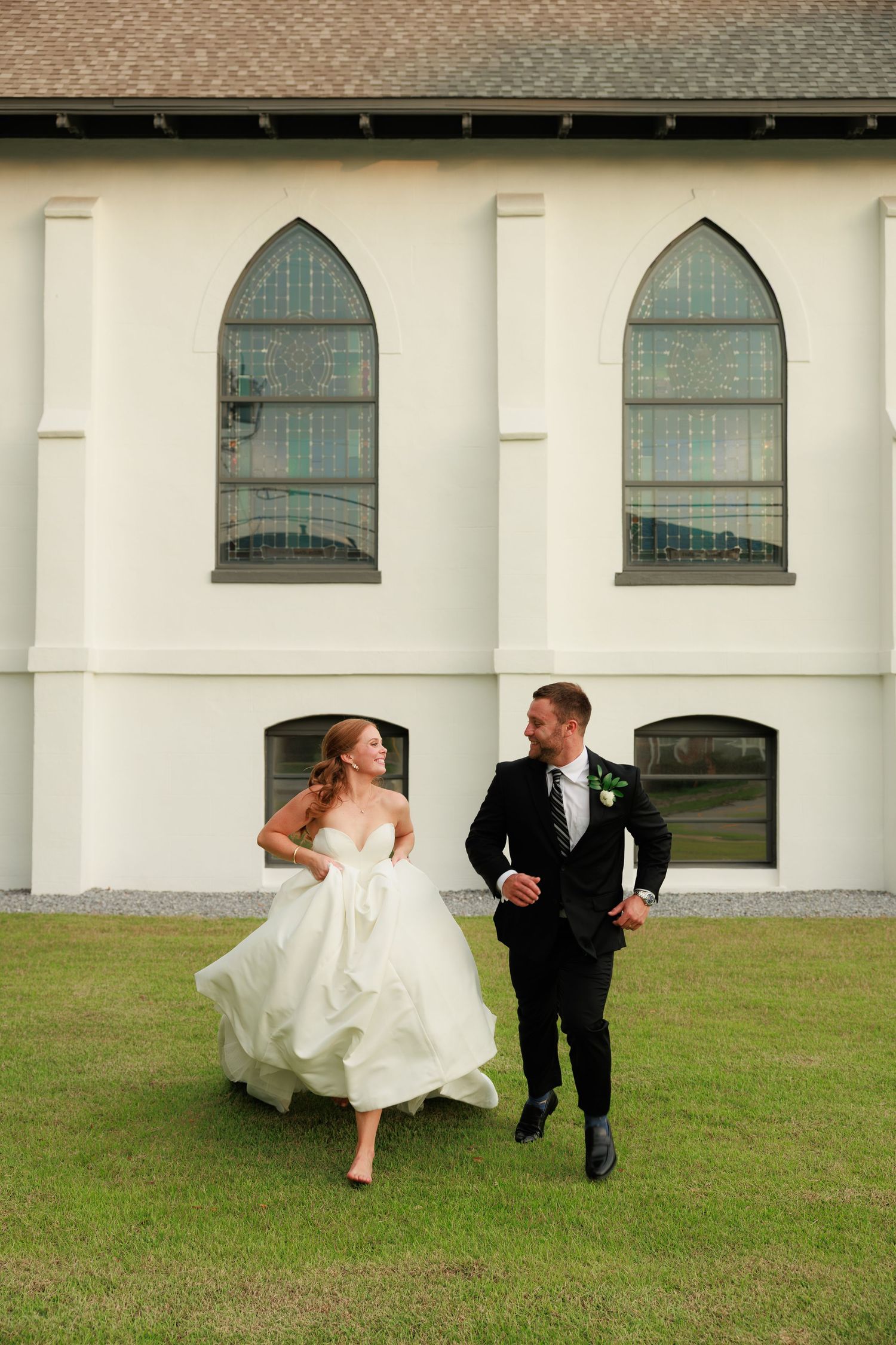 Bride and groom run joyfully across green lawn beside white church with gothic arched windows.