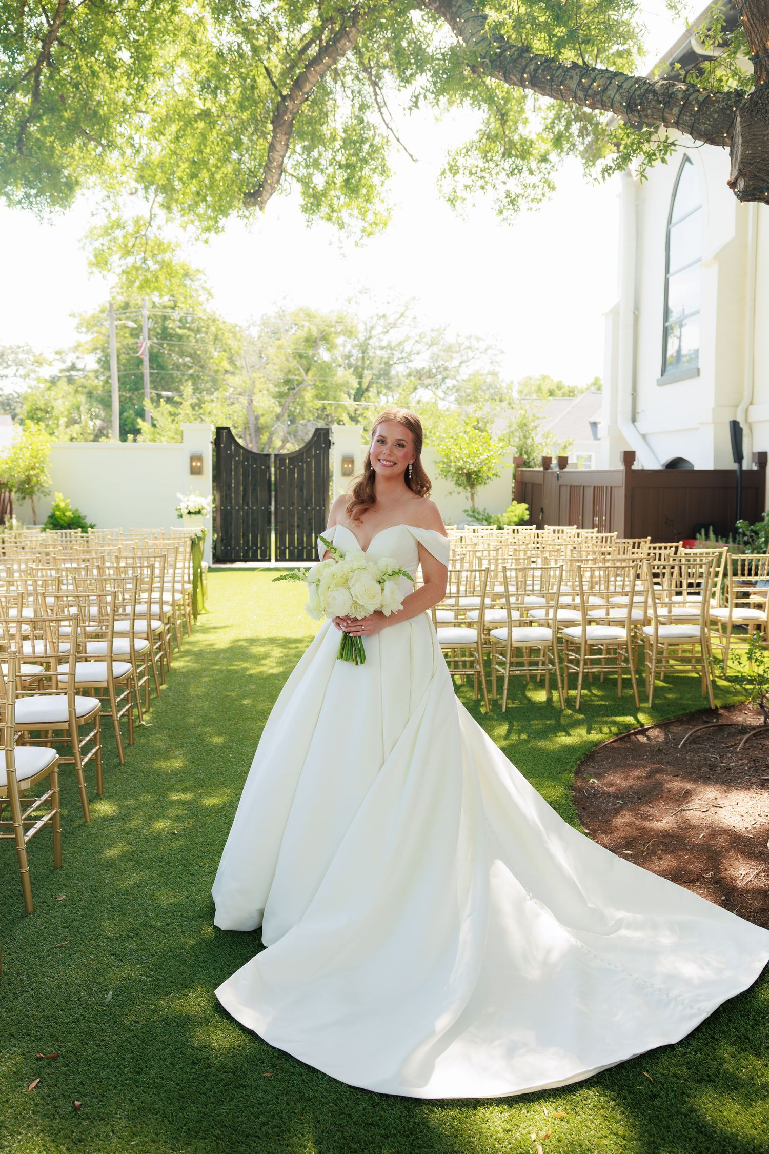 Bride in off-shoulder white ball gown holding bouquet stands on lawn with gold chairs at outdoor wedding ceremony.