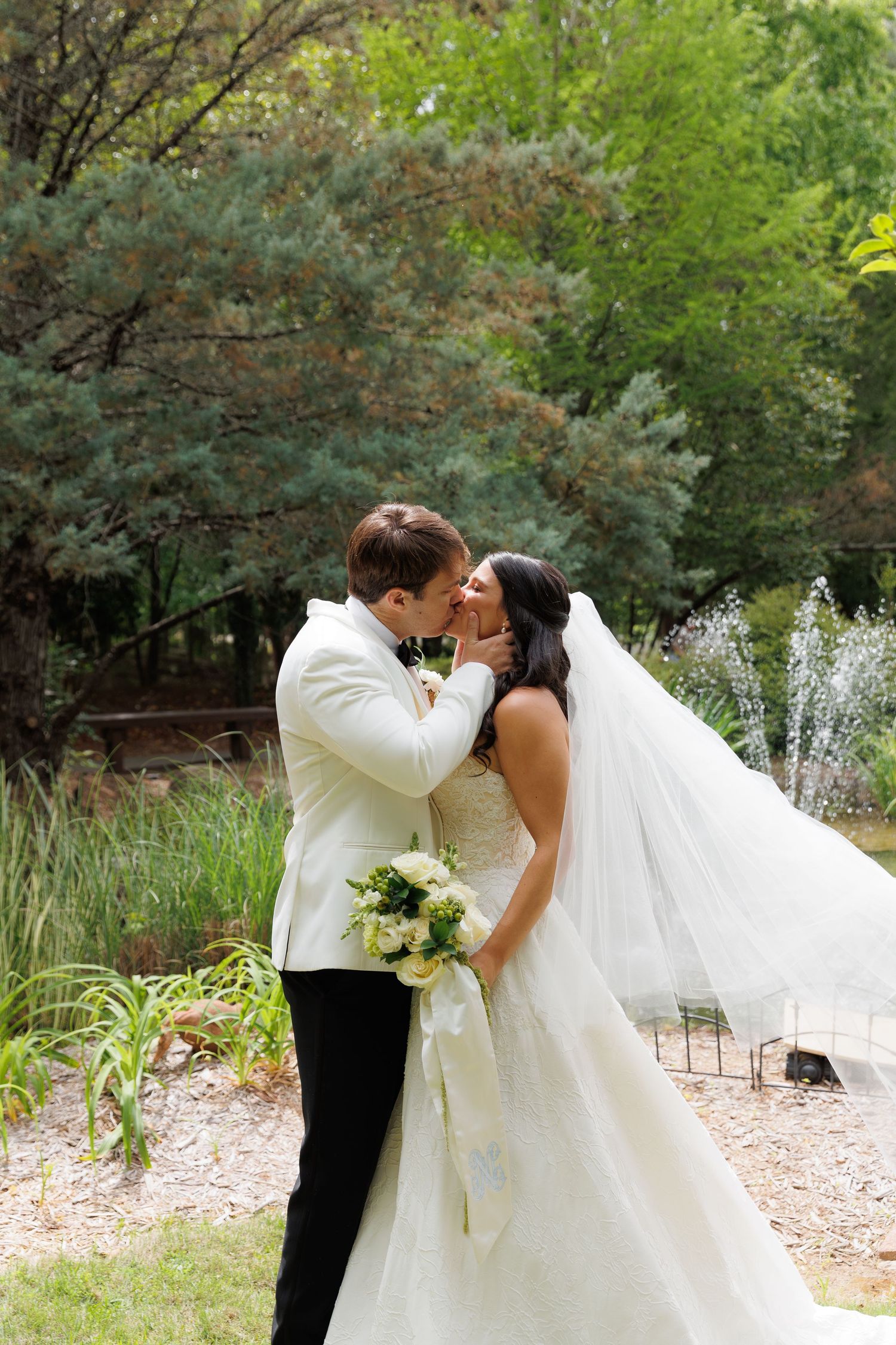 A bride and groom share a romantic kiss outdoors, with the veil flowing in the breeze near a garden fountain.