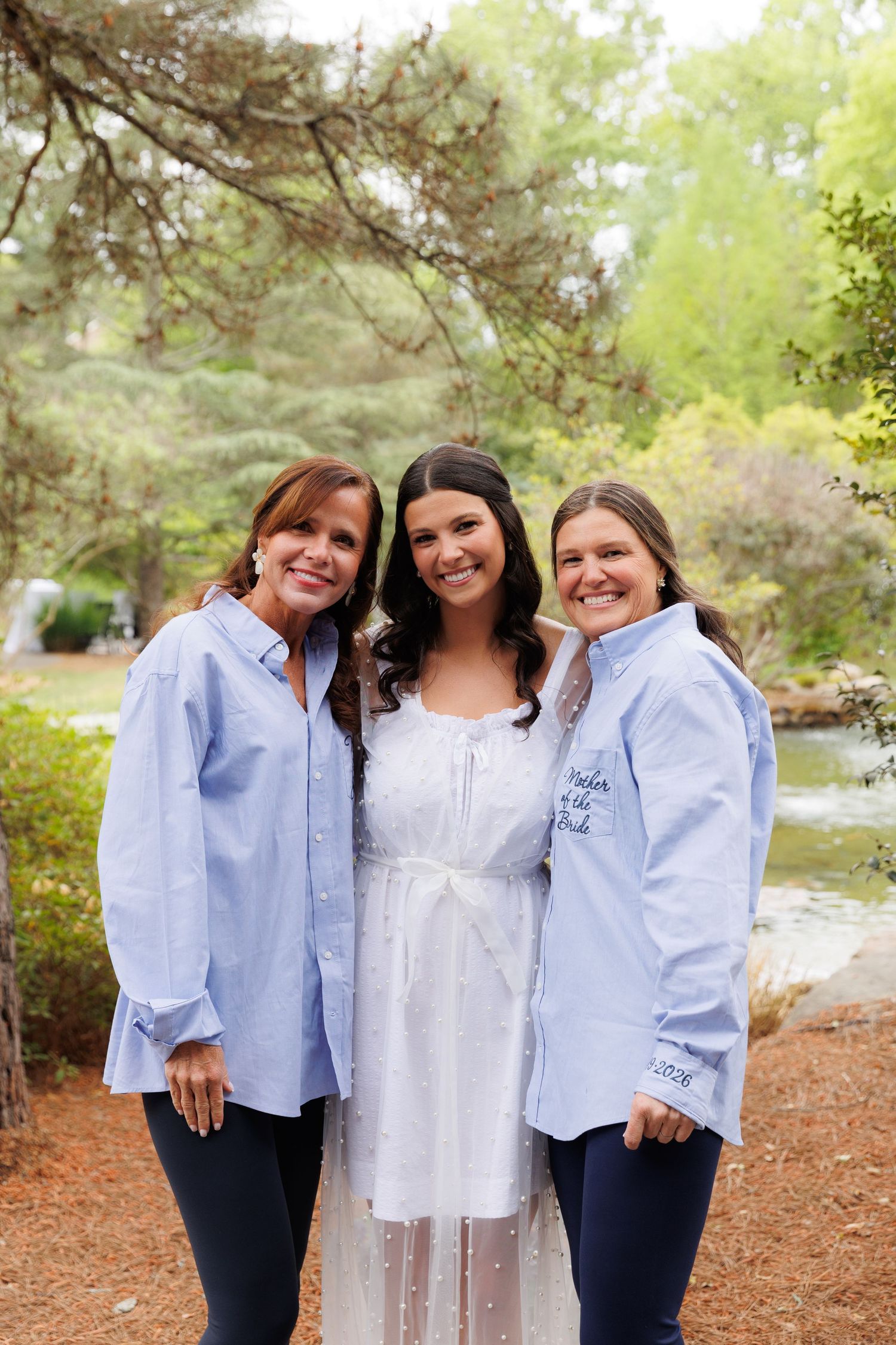Bride in white dress poses with two women in matching light blue oxford shirts in an outdoor garden setting.