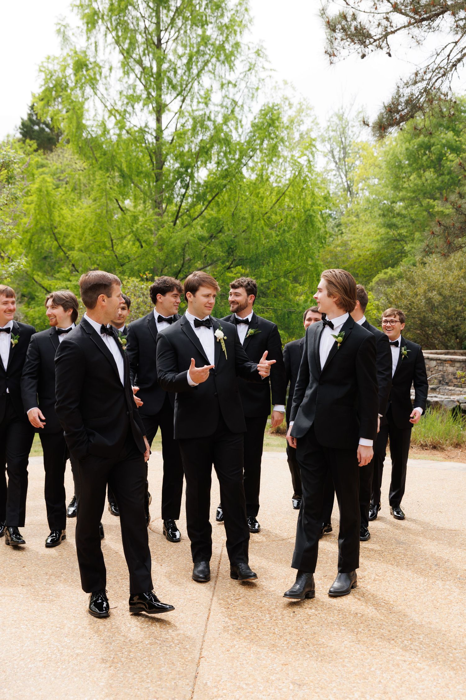 Groom and groomsmen in black tuxedos walk and chat casually on a stone patio surrounded by lush greenery.