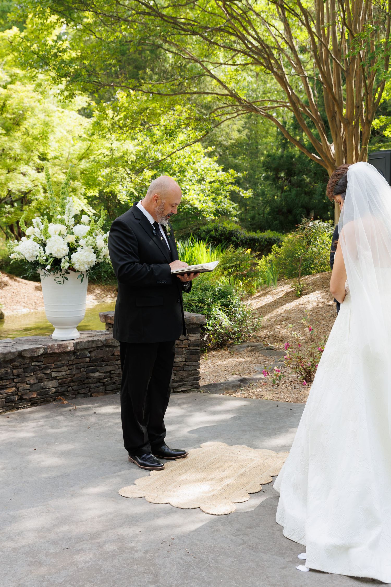 Officiant reading vows at an outdoor garden wedding ceremony with bride in white gown and veil.