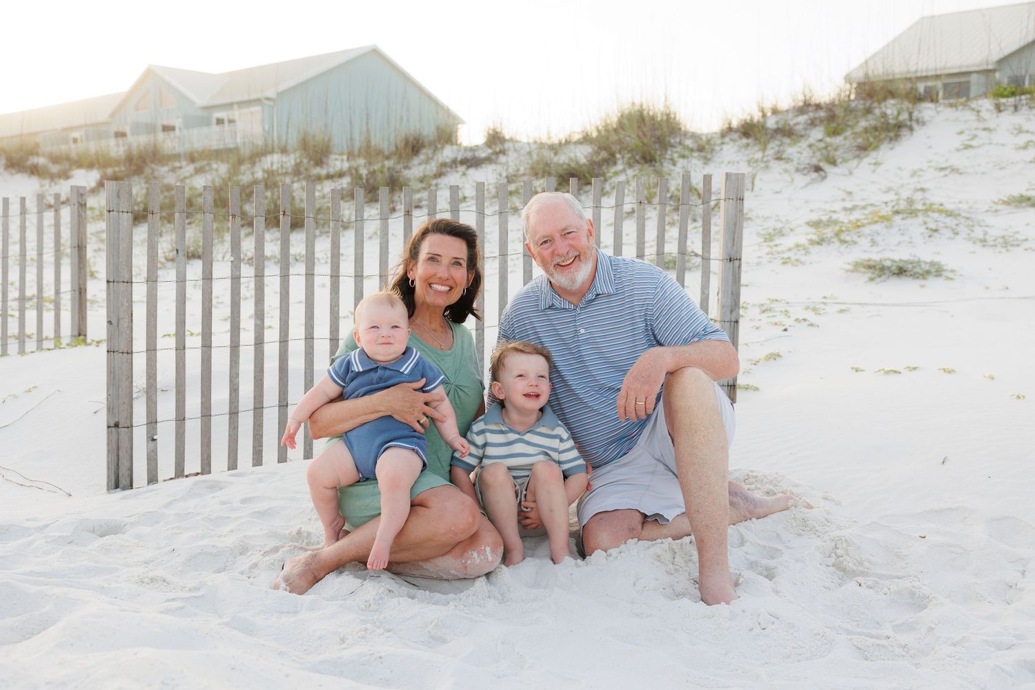 Family of four smiling on white sand beach with dunes and pastel beach houses in background on sunny day.