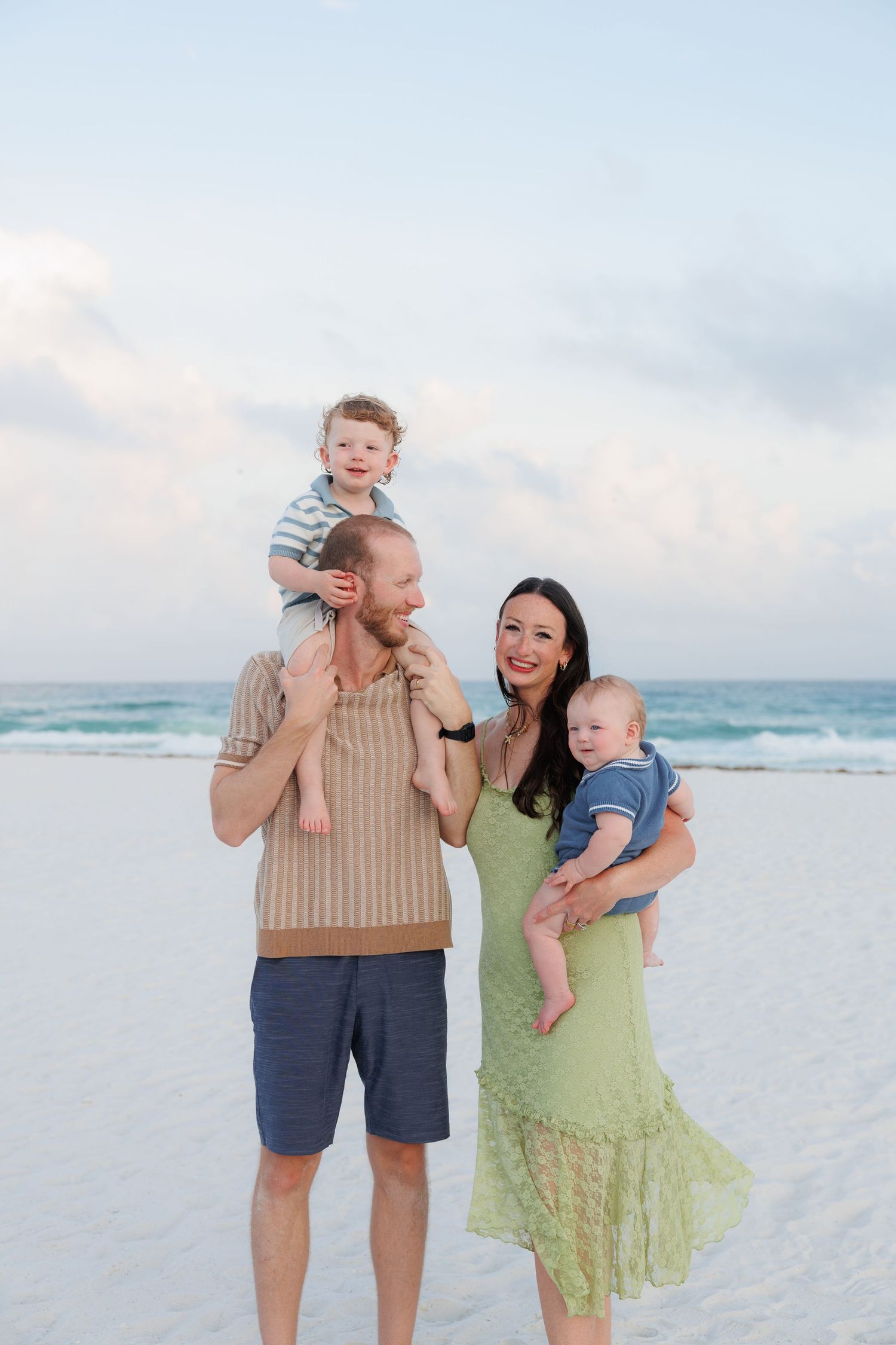 Family of four posing on a white sand beach at sunset, parents holding two young children with turquoise ocean waves behind them.
