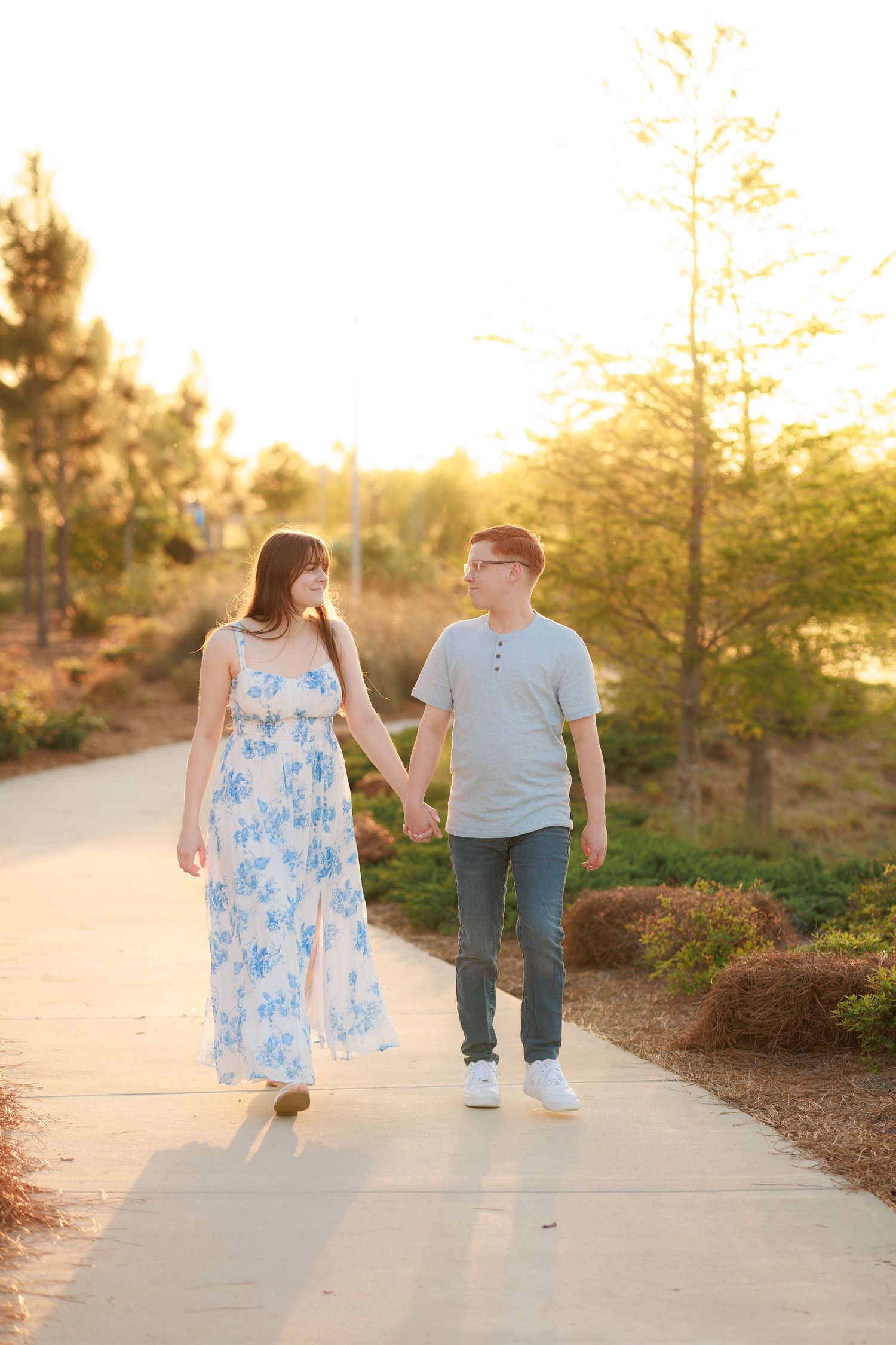 Couple walking hand in hand on a sunlit path at golden hour, woman in blue floral maxi dress, man in gray shirt.