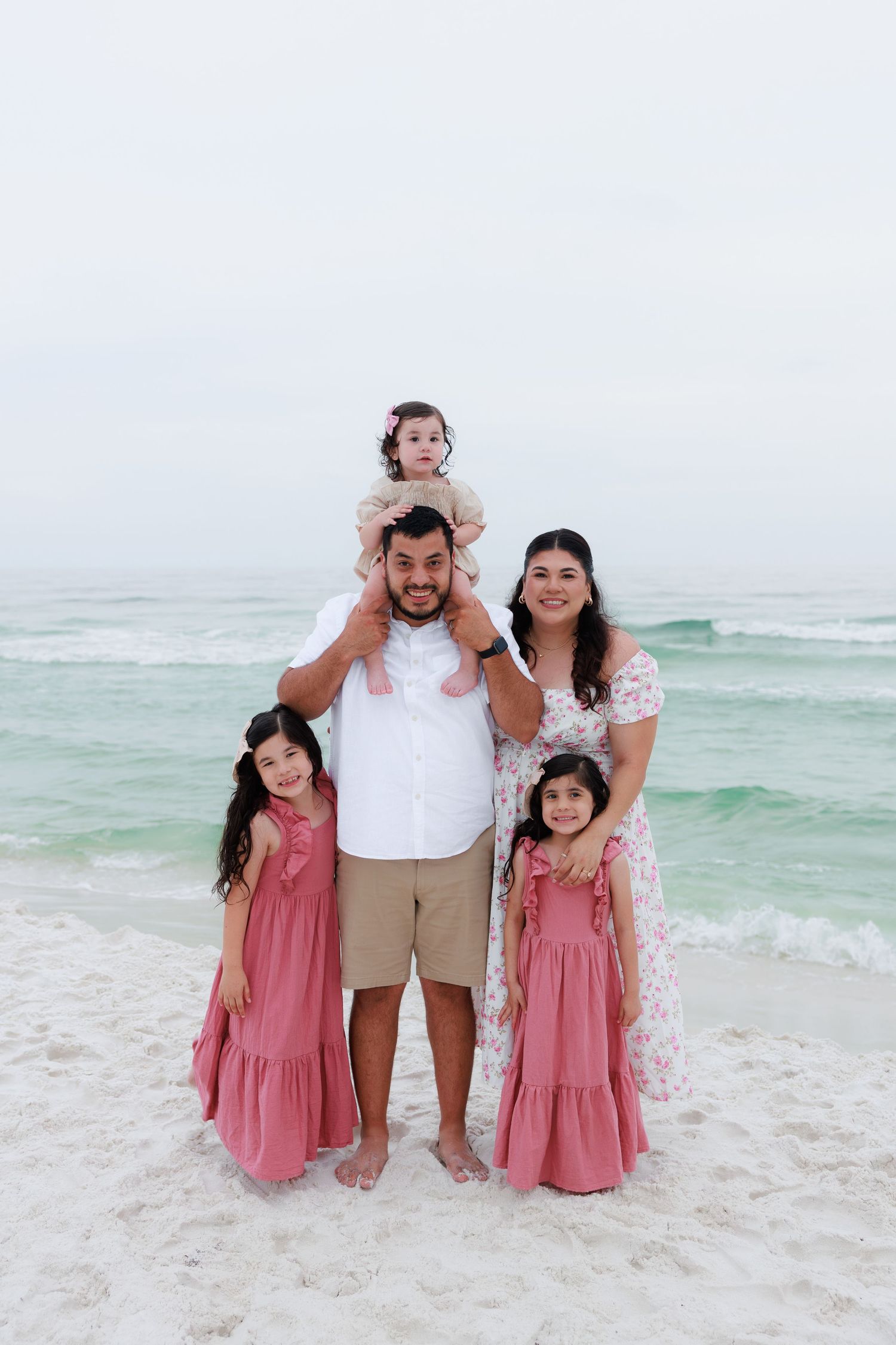 Family of five posing on a beach, parents with three young daughters in matching pink dresses, ocean waves in the background.