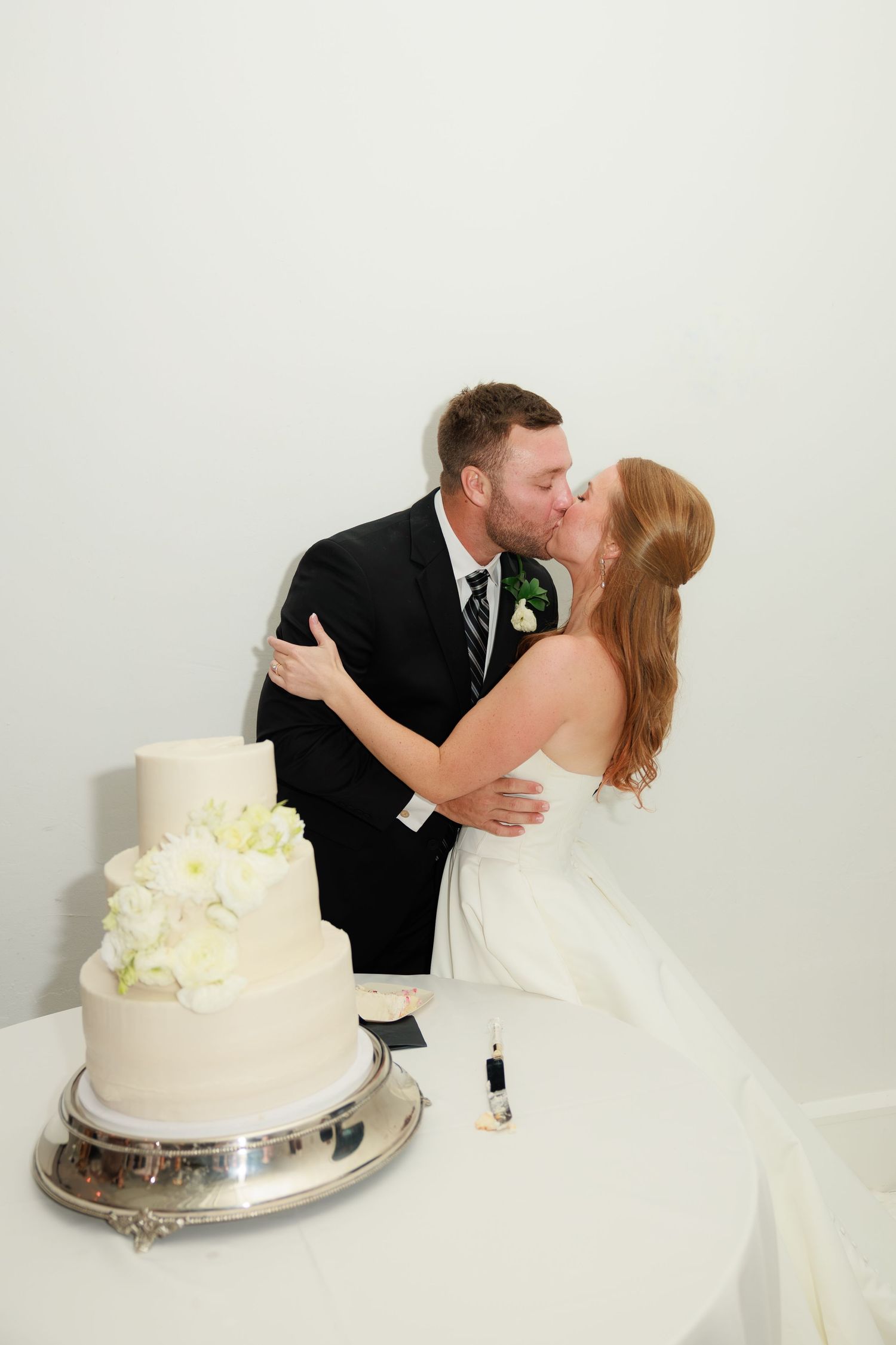Newlyweds share a kiss while cutting their white three-tier wedding cake decorated with cream flowers.