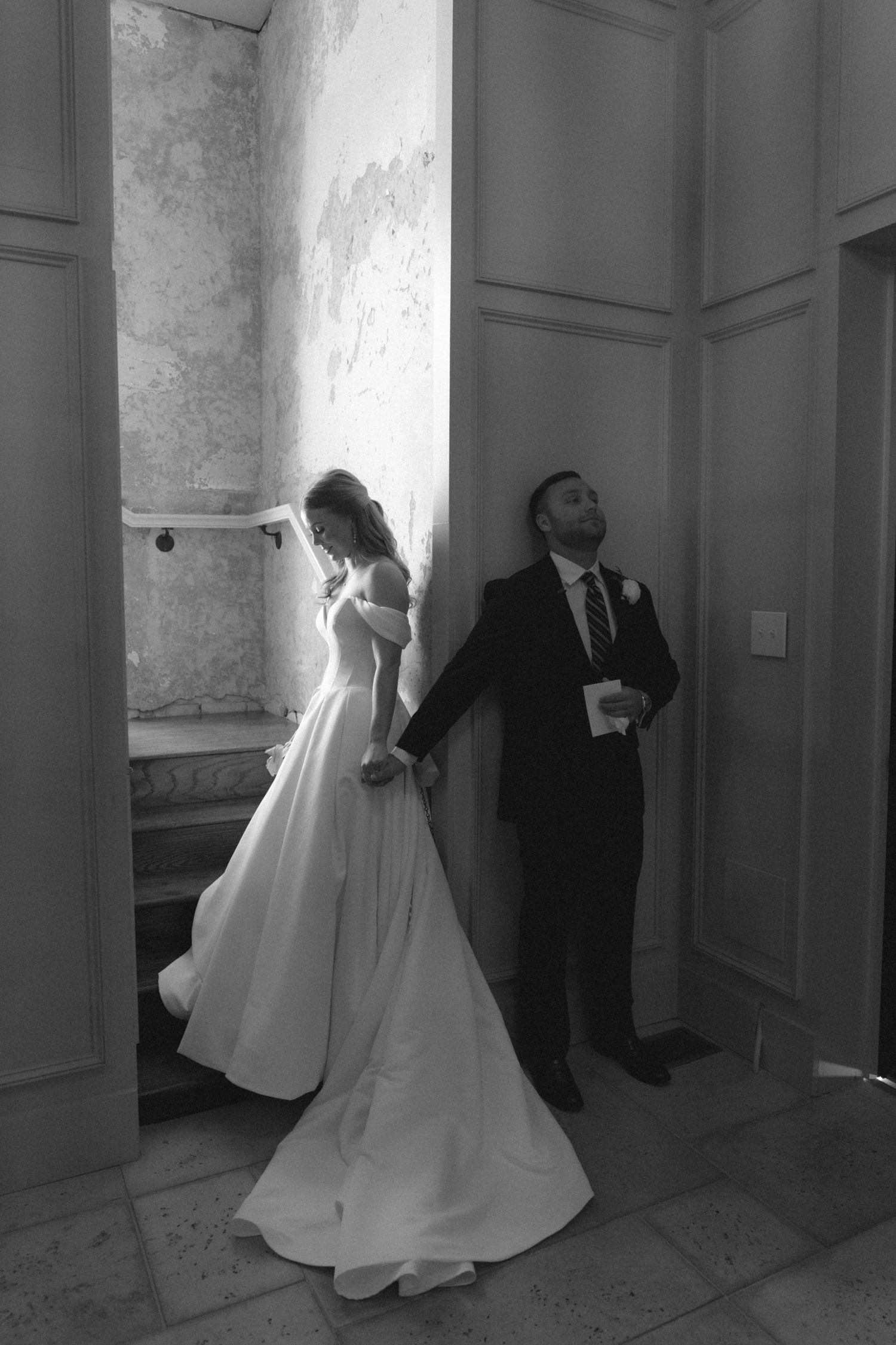 Elegant black and white wedding photo of bride and groom standing apart, bride in flowing off-shoulder gown holding hands.