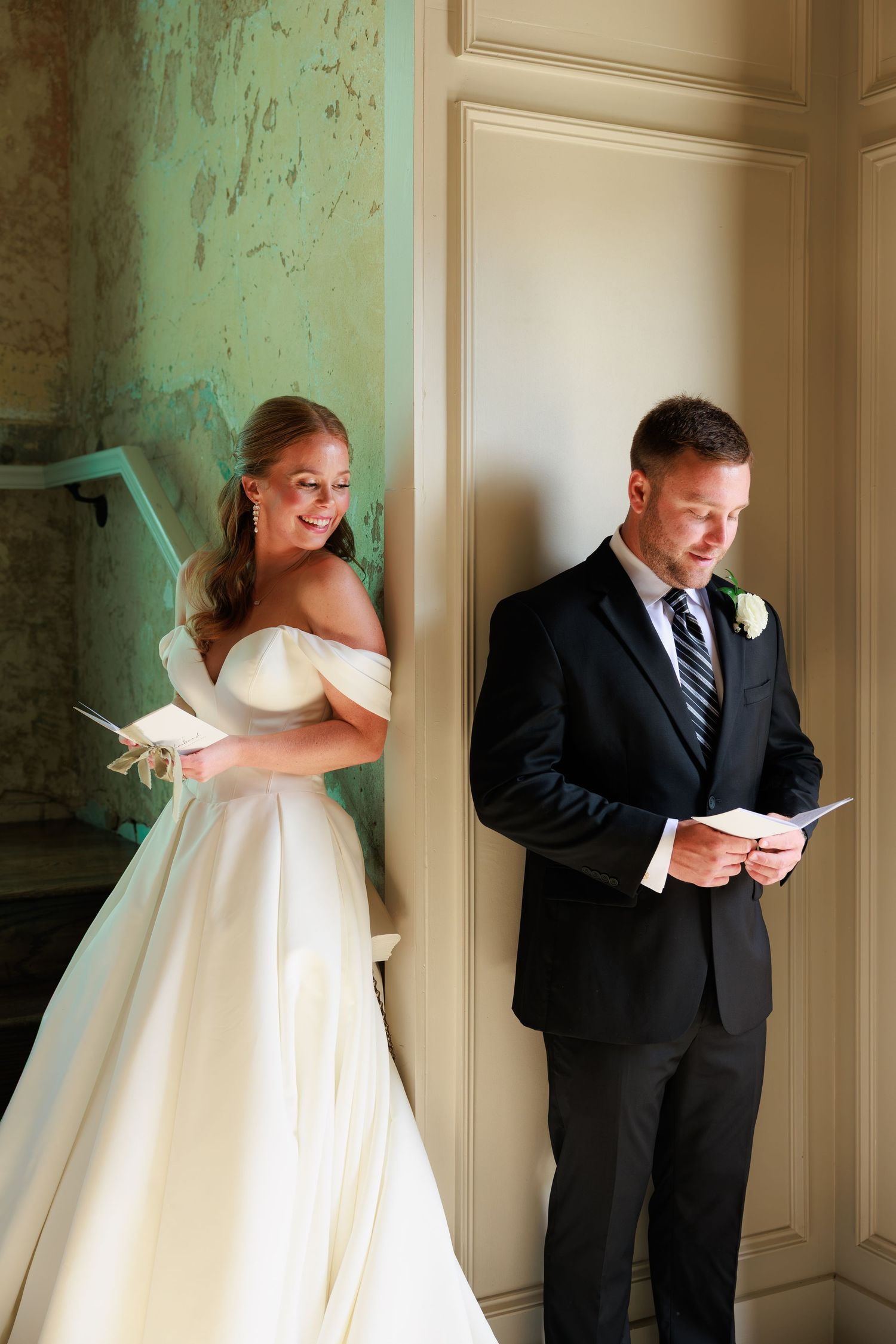 Smiling bride in satin gown watches groom read wedding vow letter in a sunlit elegant hall.
