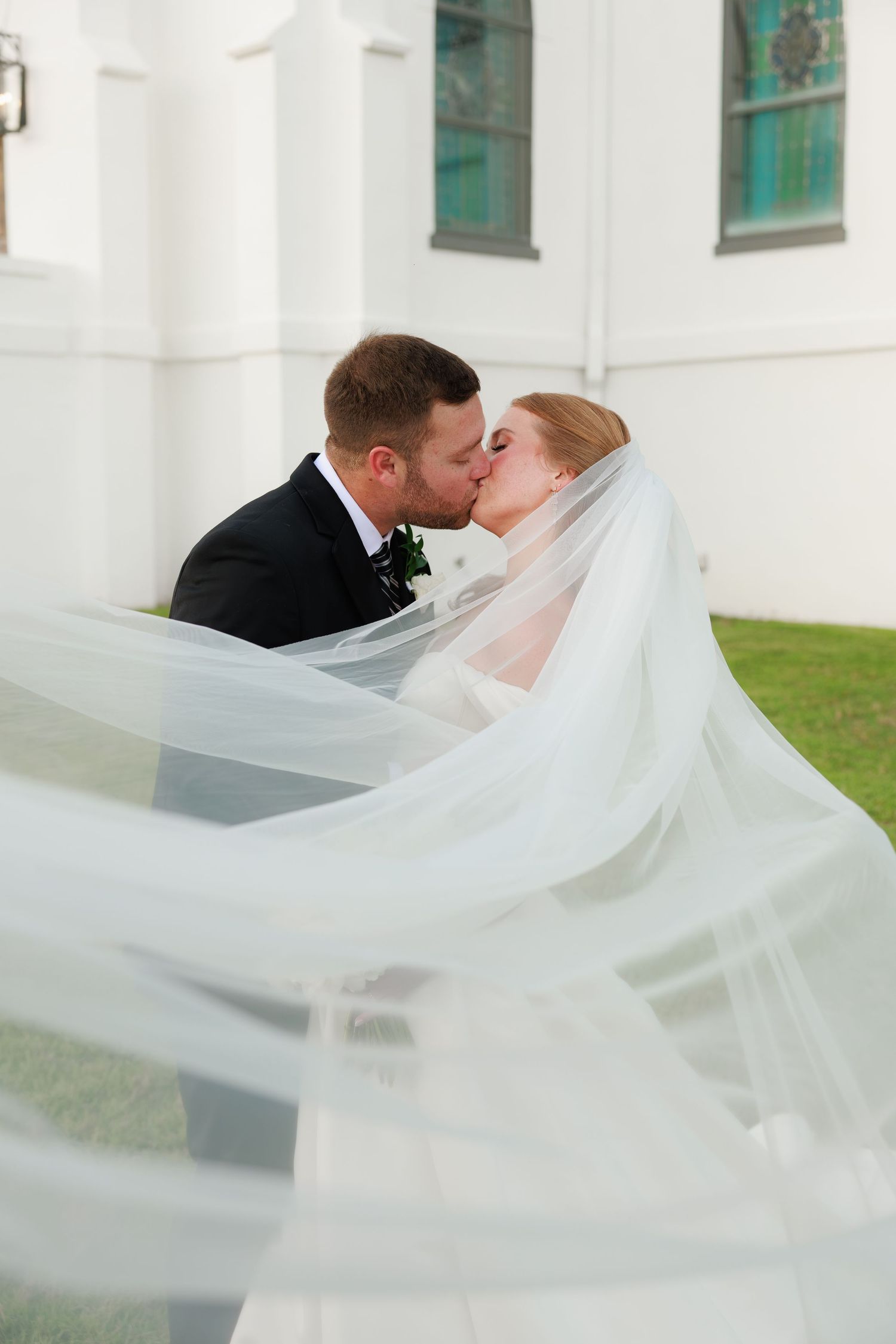 Bride and groom share a kiss as cathedral-length veil billows dramatically in the wind outside white church.