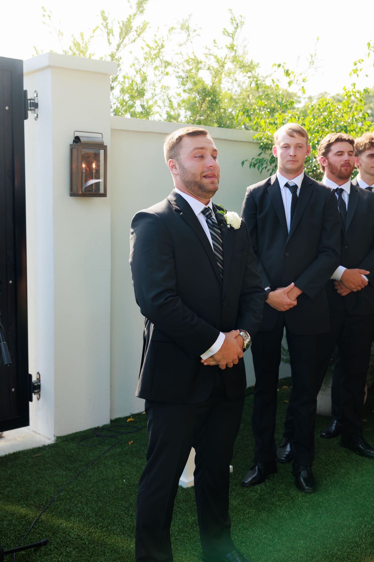 Groom in black tuxedo stands with groomsmen at outdoor wedding ceremony.