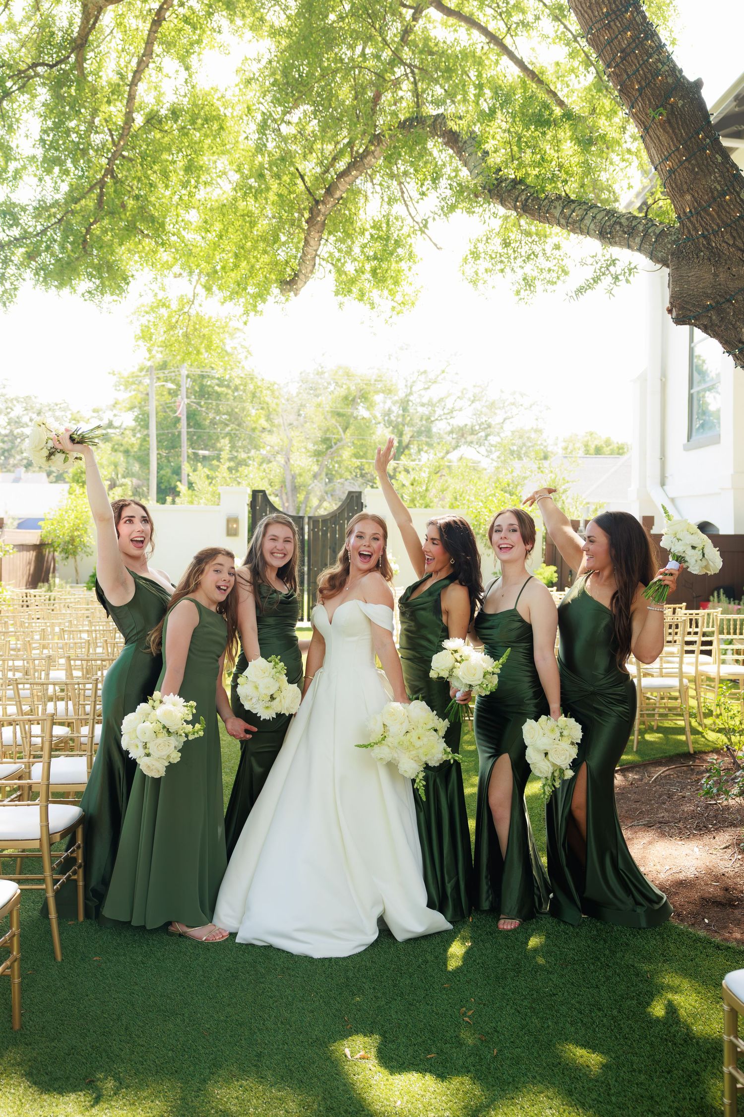 Bride in white gown celebrates with bridesmaids in olive green dresses holding white bouquets under a tree.