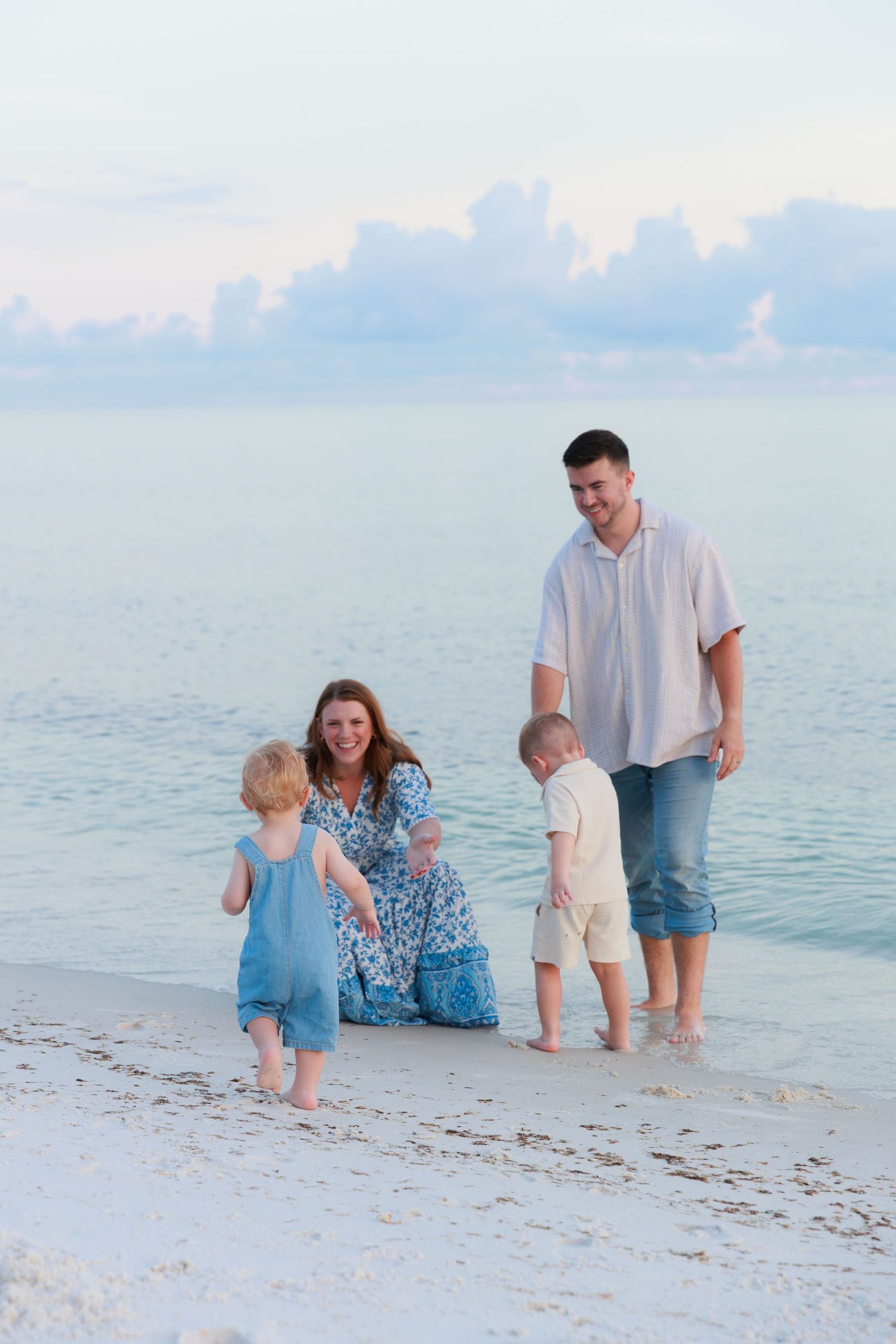A happy family of four plays together at the water's edge on a white sand beach at dusk.