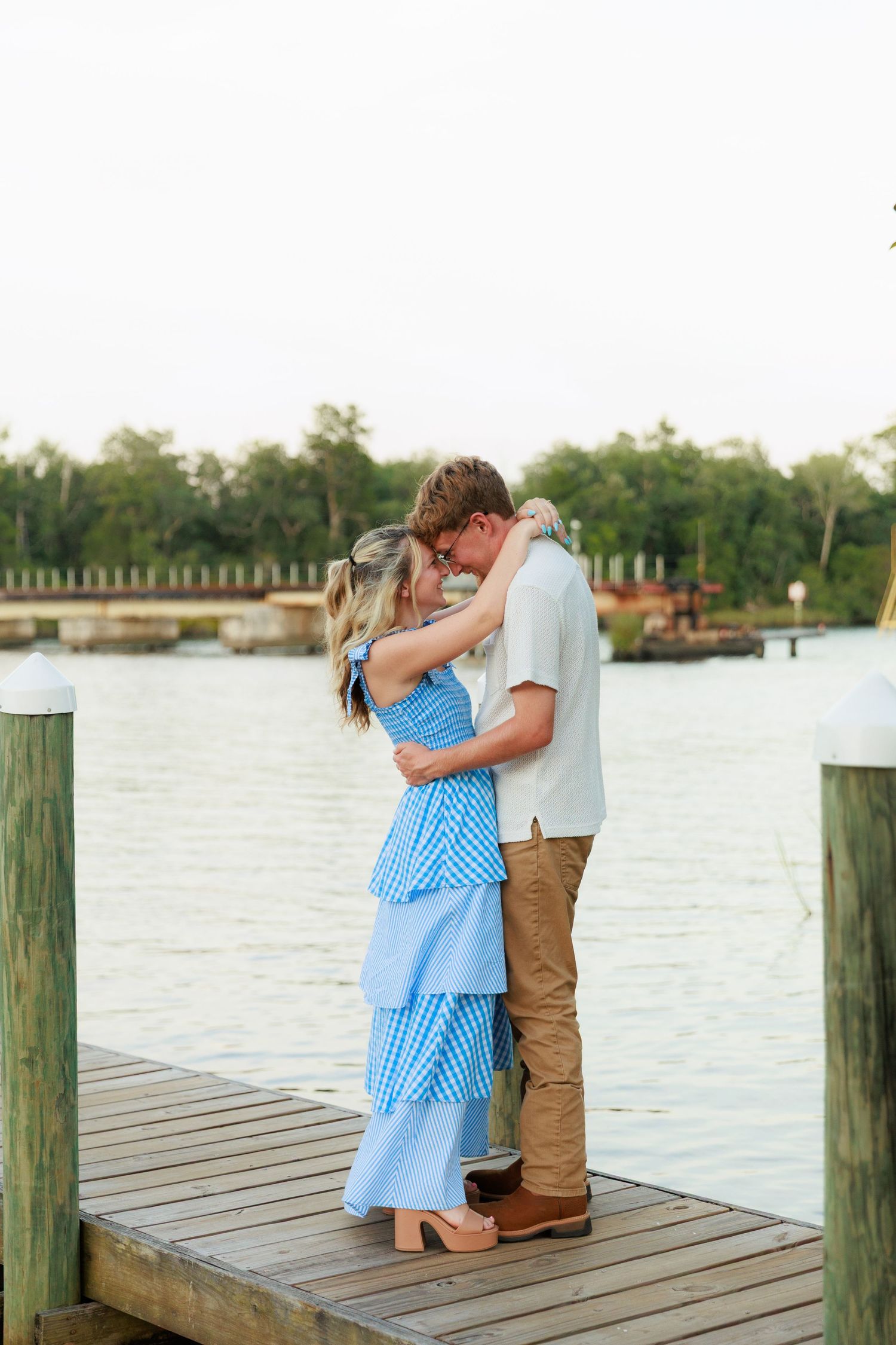 An engaged couple embraces closely on a wooden dock with a calm waterway and trees visible in the background.