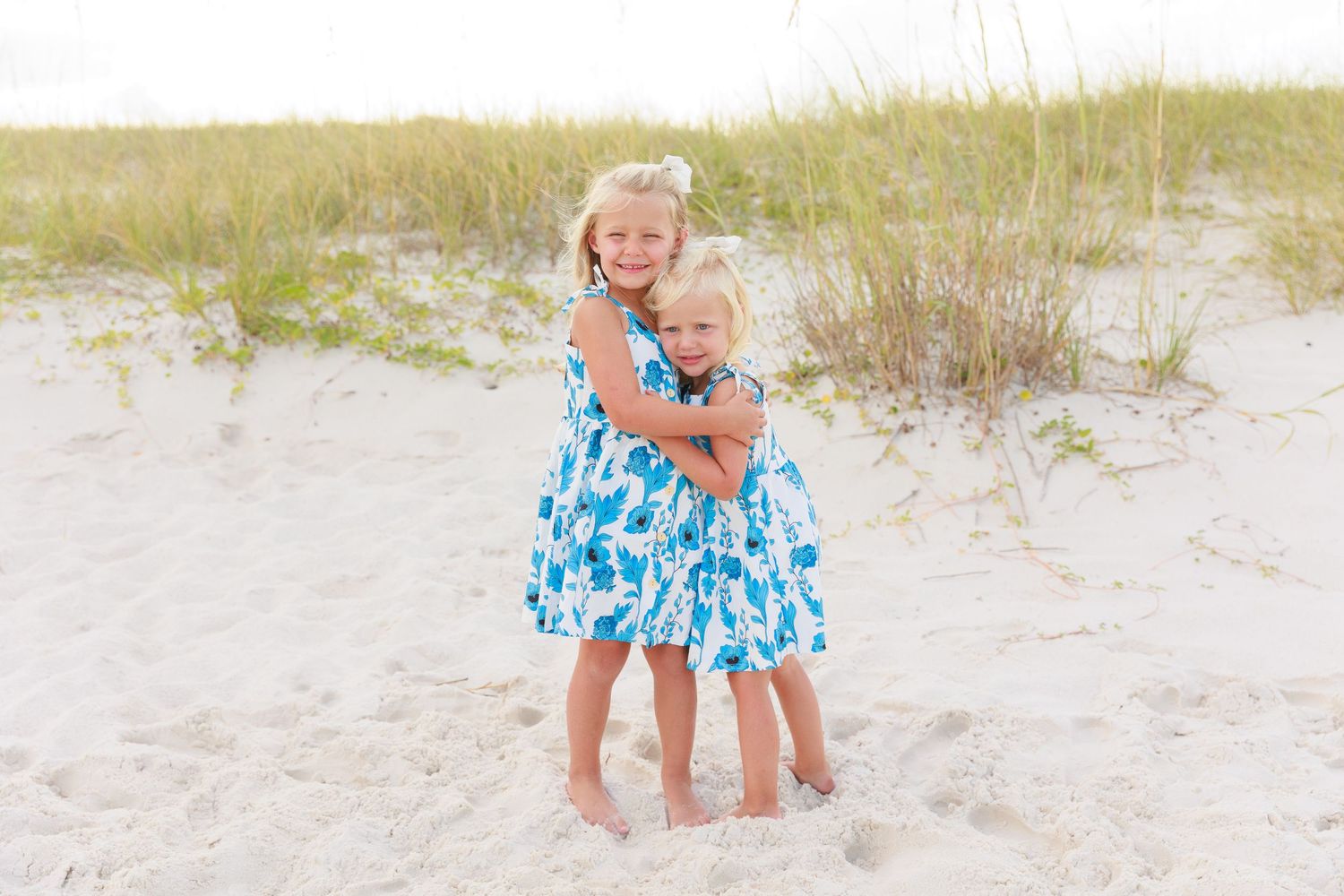 Two young girls in matching blue floral dresses hugging on a sandy beach with dunes and grass in the background.