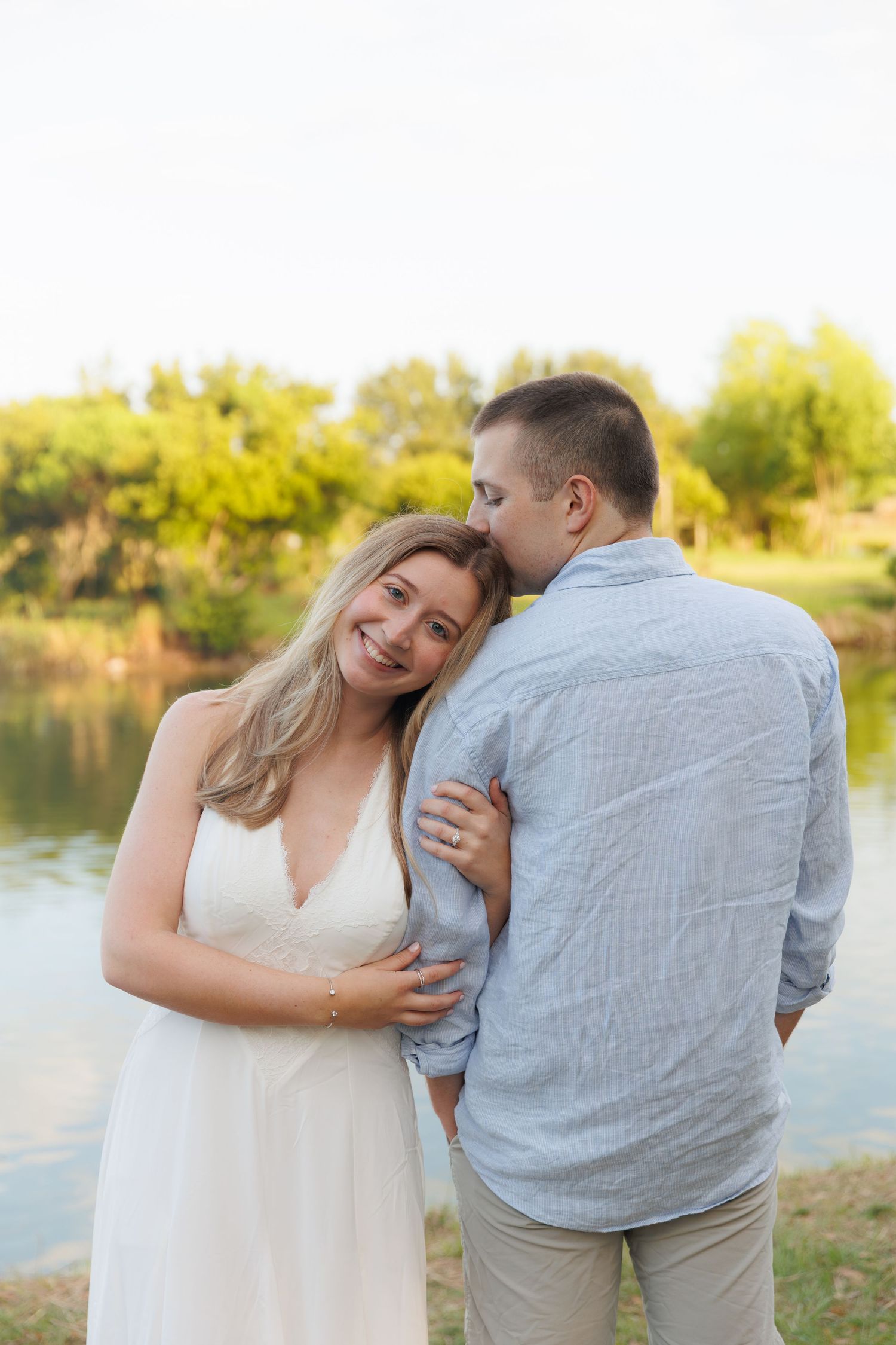 Couple sharing a romantic kiss by a serene lake, woman in white dress smiling, golden hour lighting in lush park.
