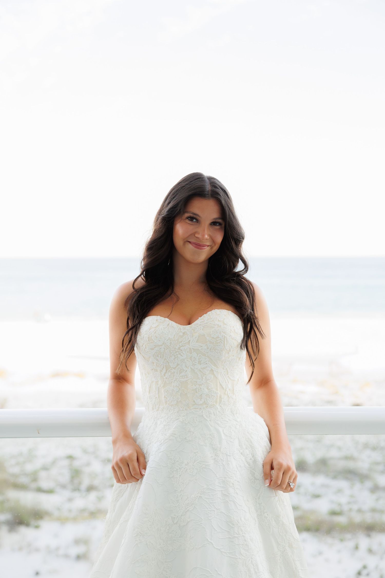 Beautiful bride in strapless lace wedding gown poses on beach balcony.