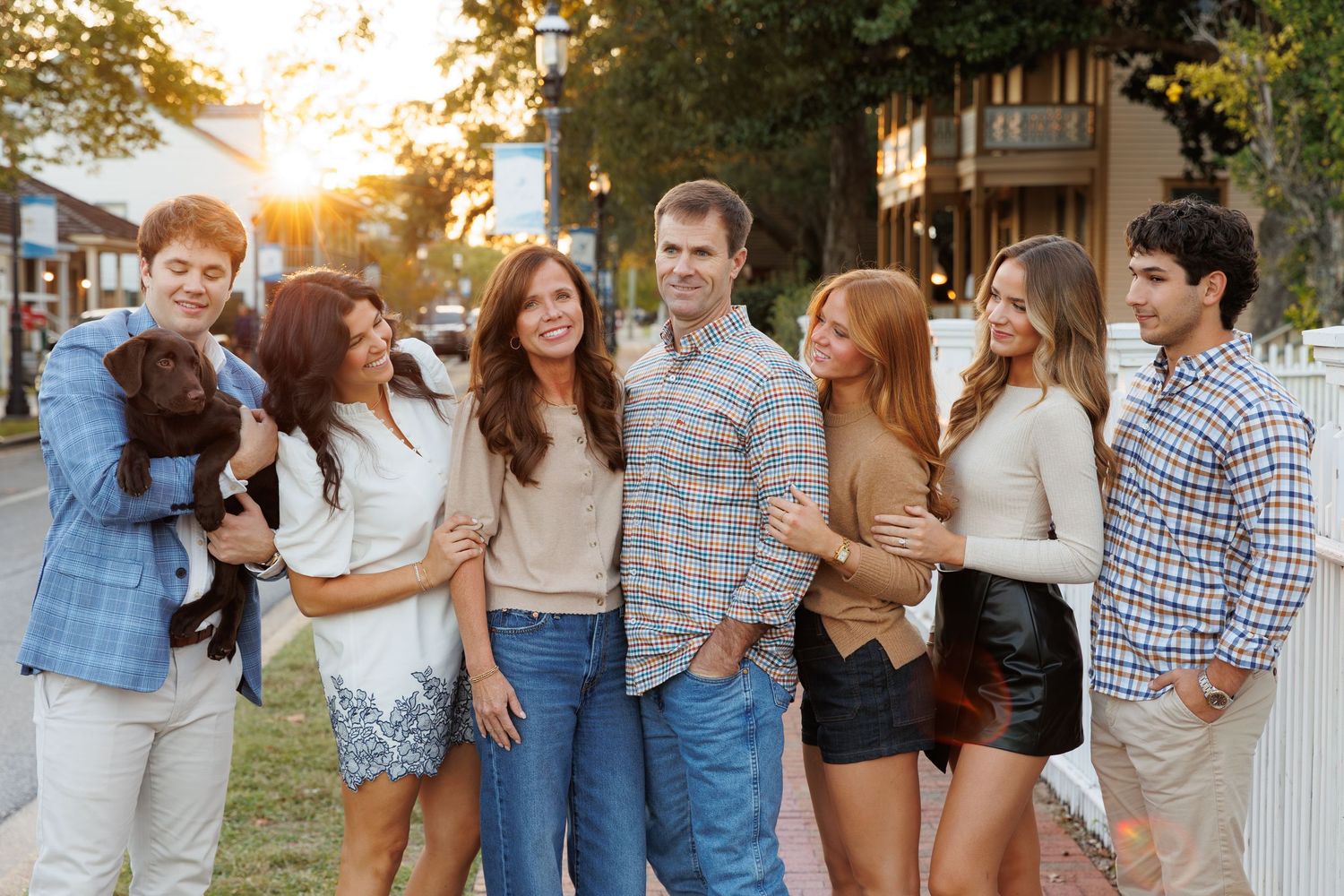 A cheerful family group poses outdoors at golden hour, with a young man holding a puppy on the left.