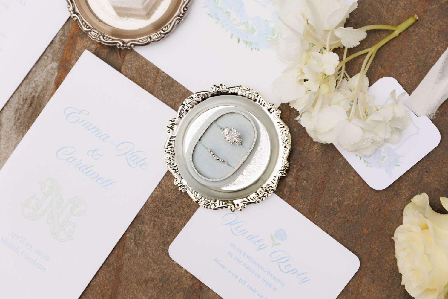 Elegant wedding stationery flat lay with silver ornate mirror, diamond ring, and white floral accents on rustic wood.