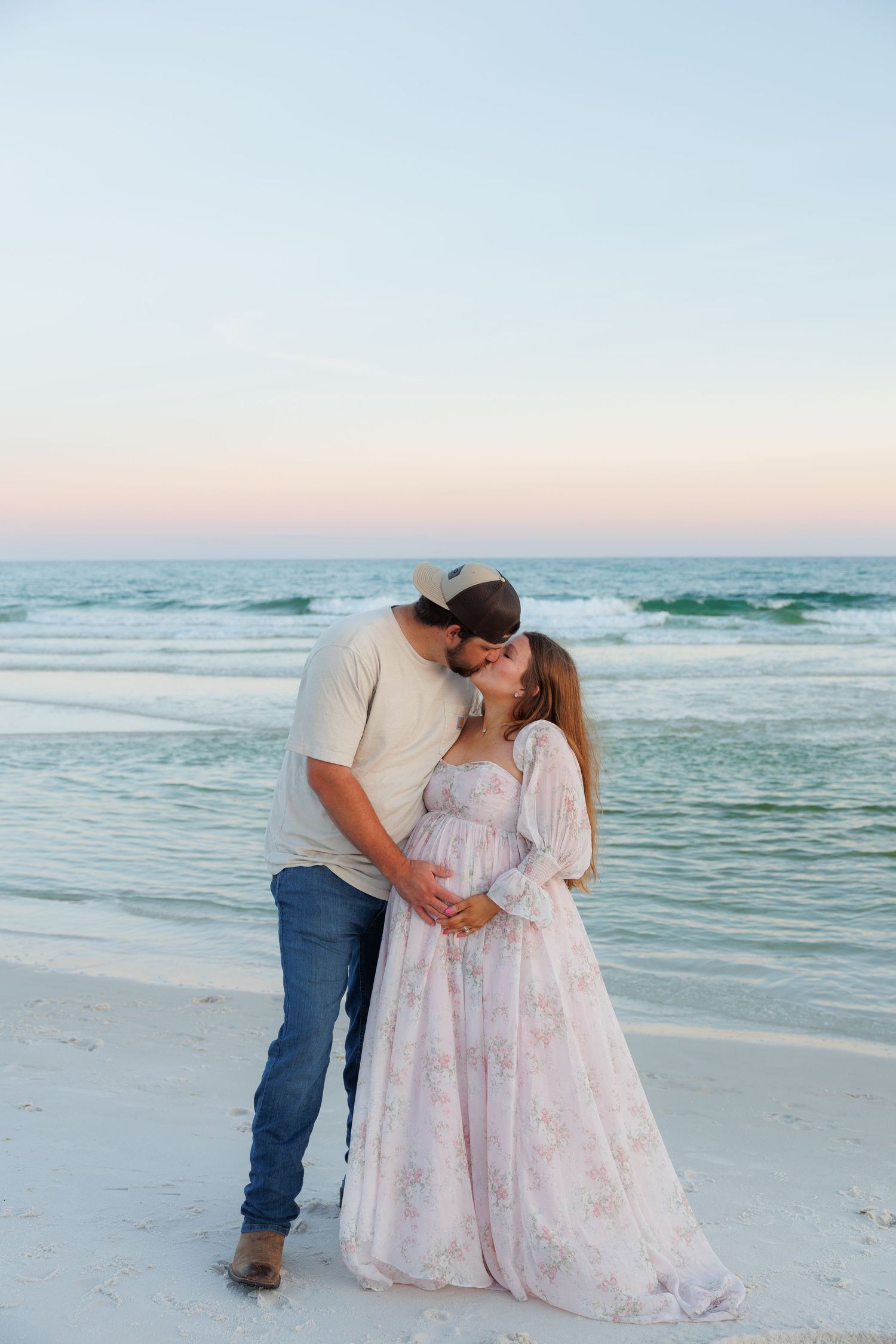 Couple sharing a kiss on a white sand beach at sunset, woman in floral maternity gown cradling baby bump.
