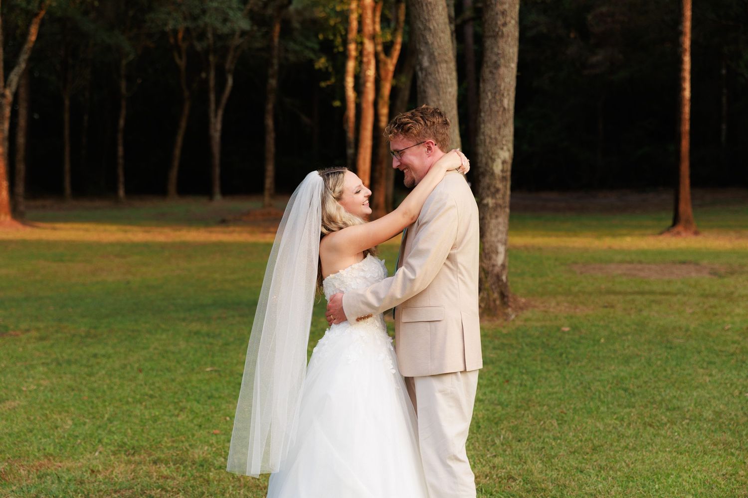 A couple in wedding attire shares a romantic moment at sunset in a wooded outdoor setting.