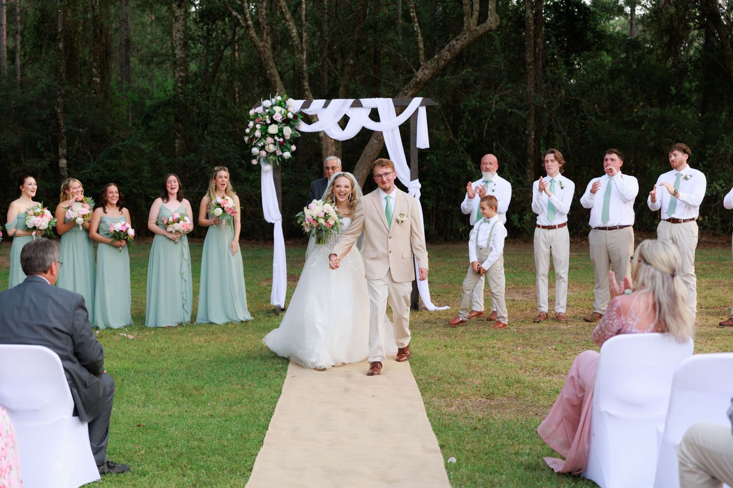 A wedding party in white and blush pink attire poses together on a grassy lawn during an outdoor celebration.