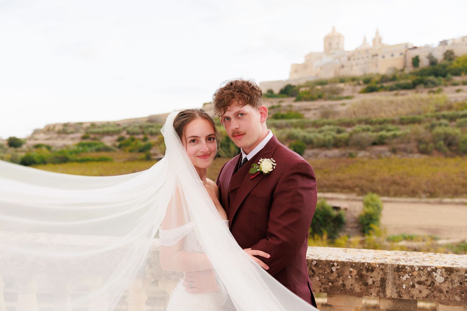 Newlywed couple embraces on a scenic hilltop overlooking a rustic countryside landscape with stone walls.