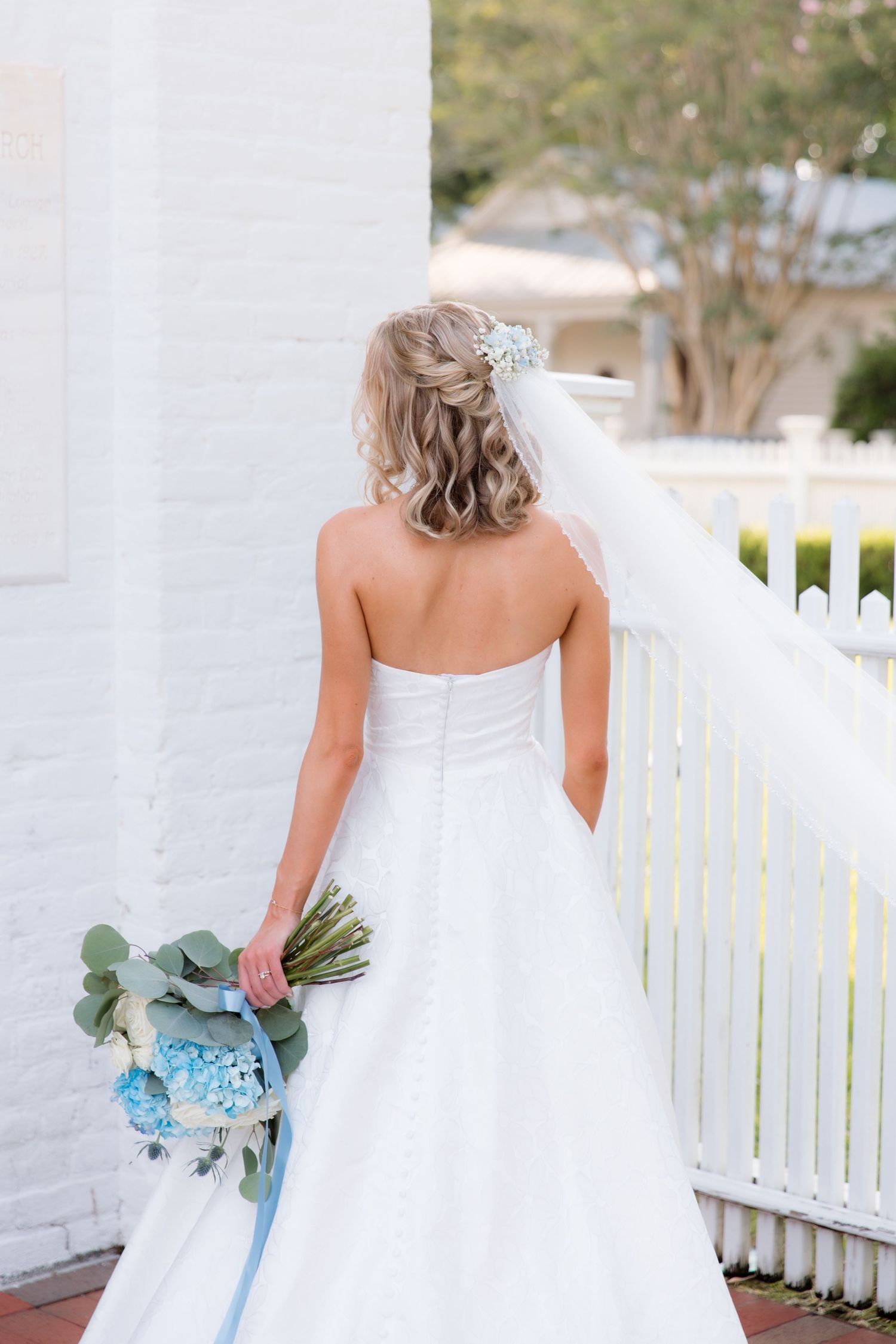 Back view of bride in strapless gown holding blue and white bouquet on white porch.
