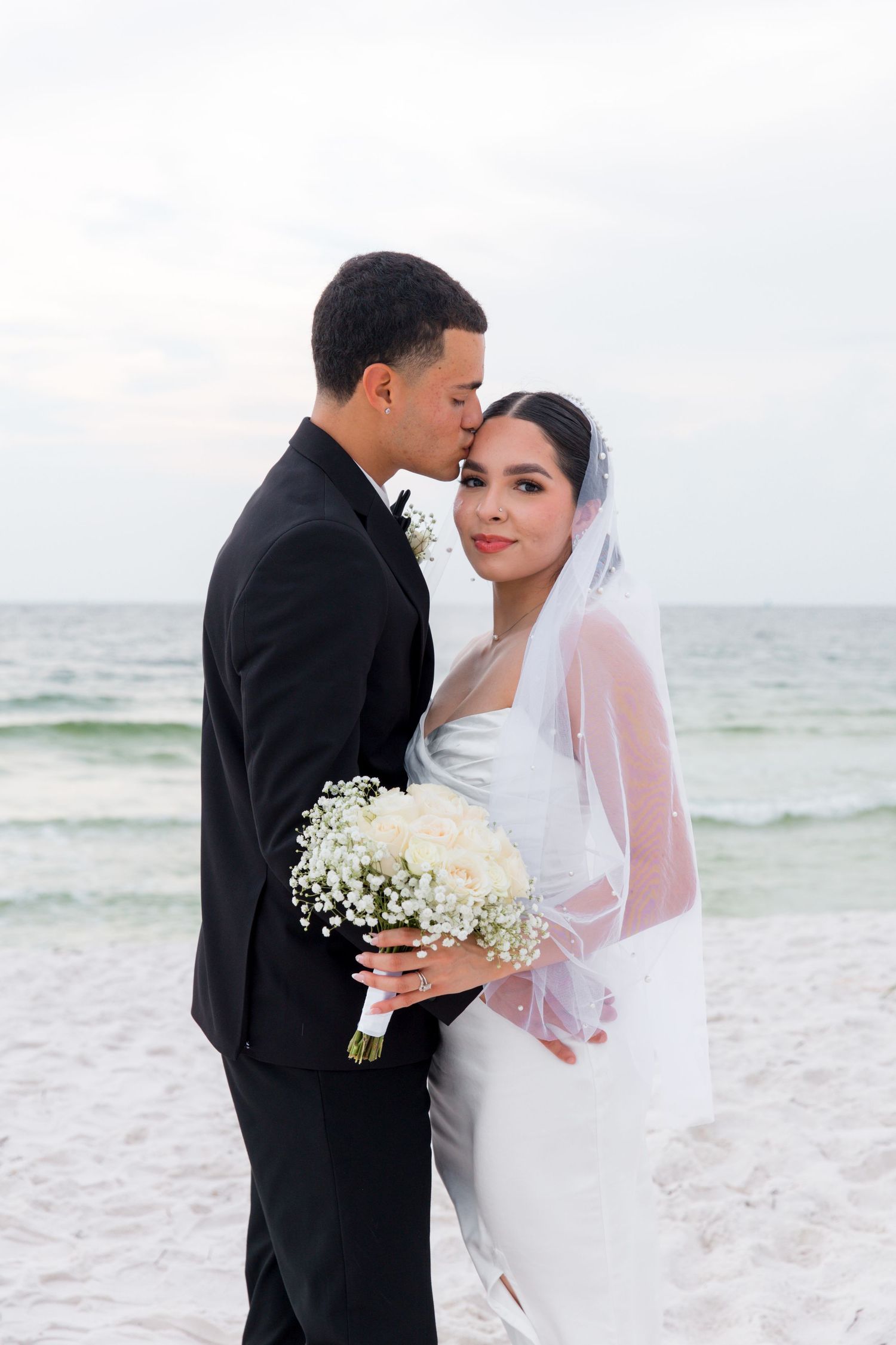 Couple shares tender moment on beach with bride holding elegant white floral bouquet.