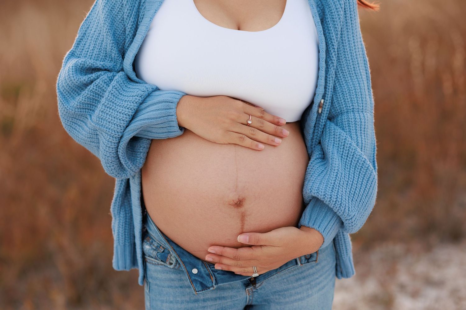 Close up of pregnant belly with hands cradling bump, wearing blue cardigan and distressed jeans.