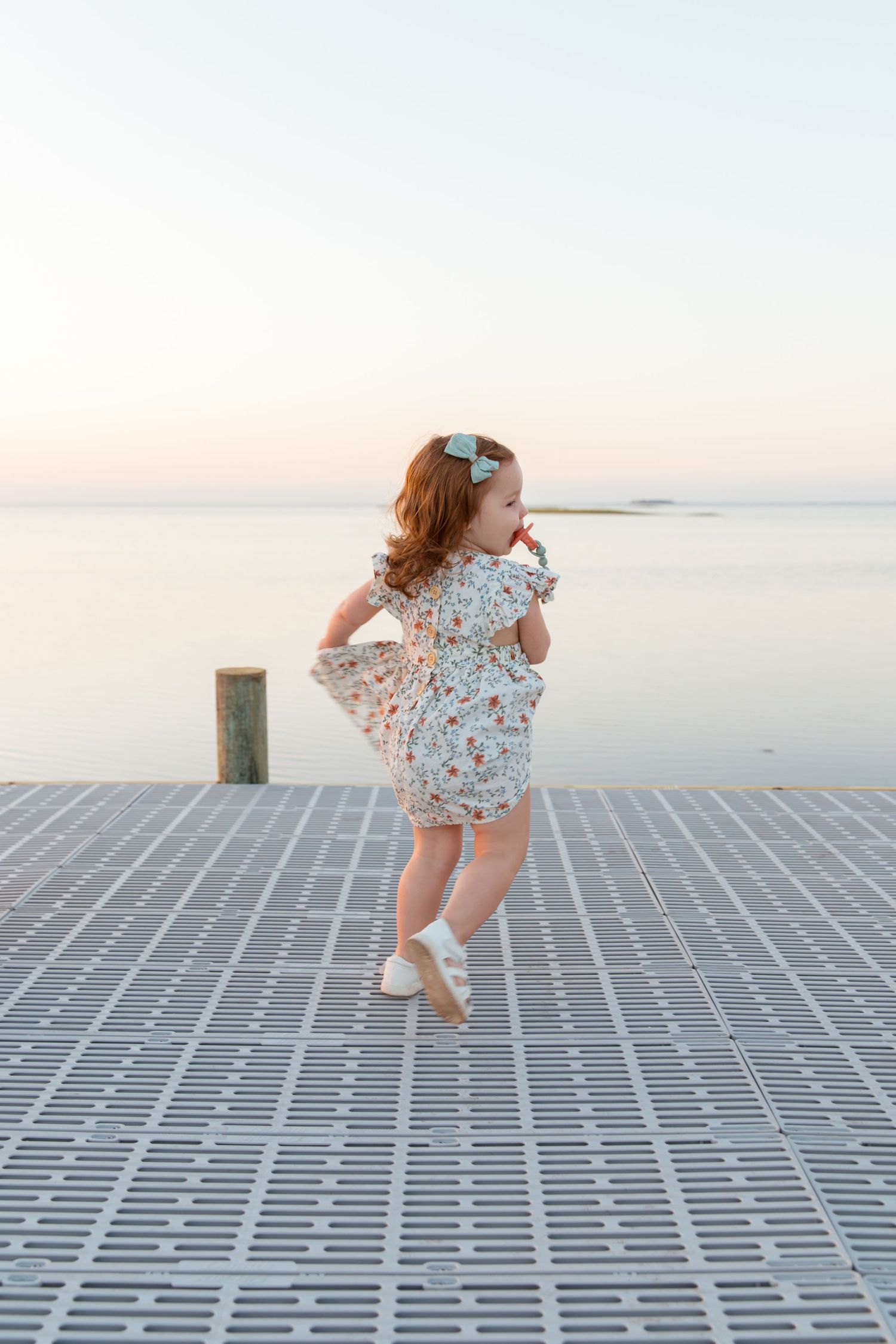 Young child in floral dress dances and twirls joyfully on wooden boardwalk at sunset.