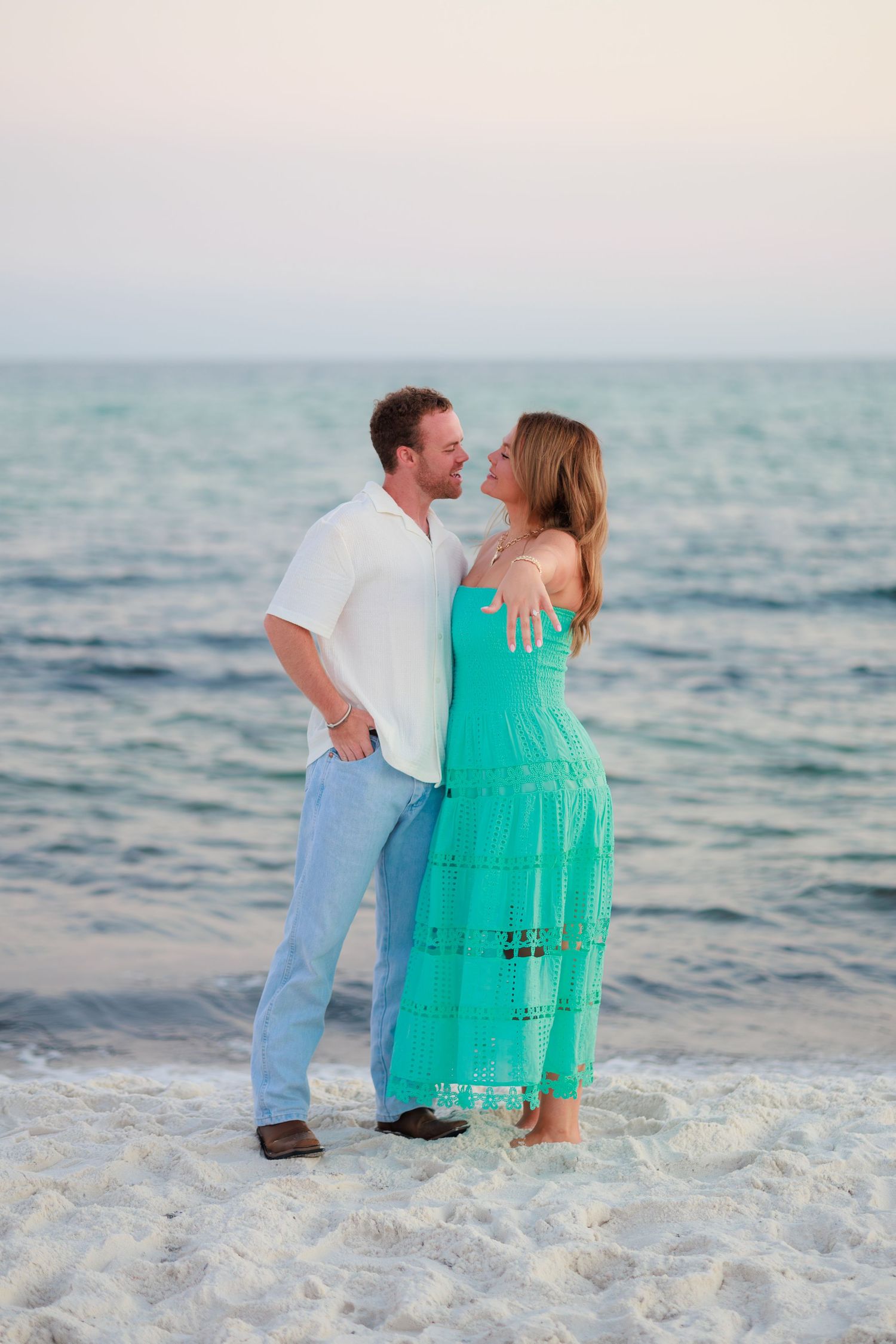A couple in casual beach attire embraces while standing on white sand with turquoise ocean waters in the background.
