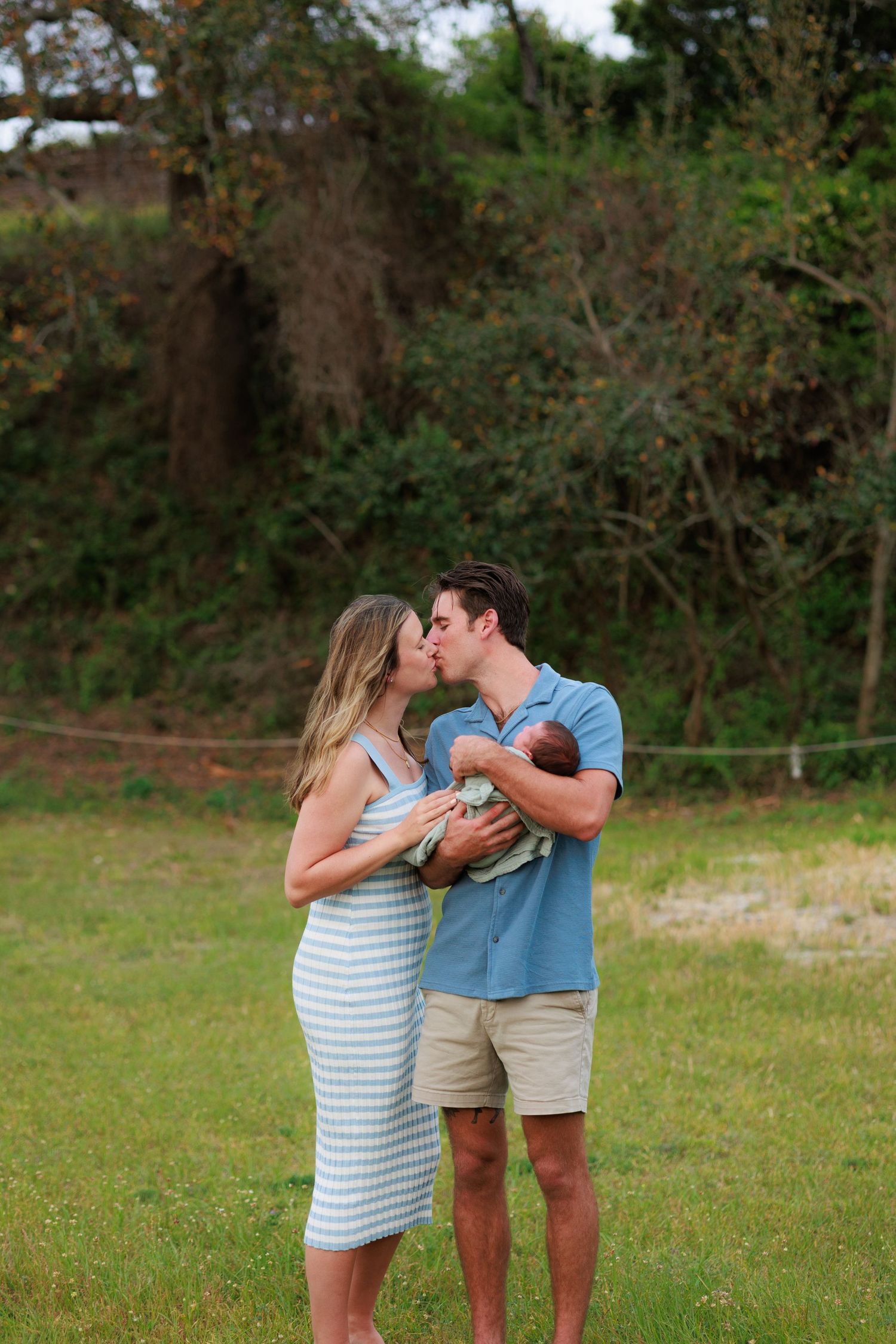 A couple shares a drink from bottles while standing together in a grassy field on a sunny day.