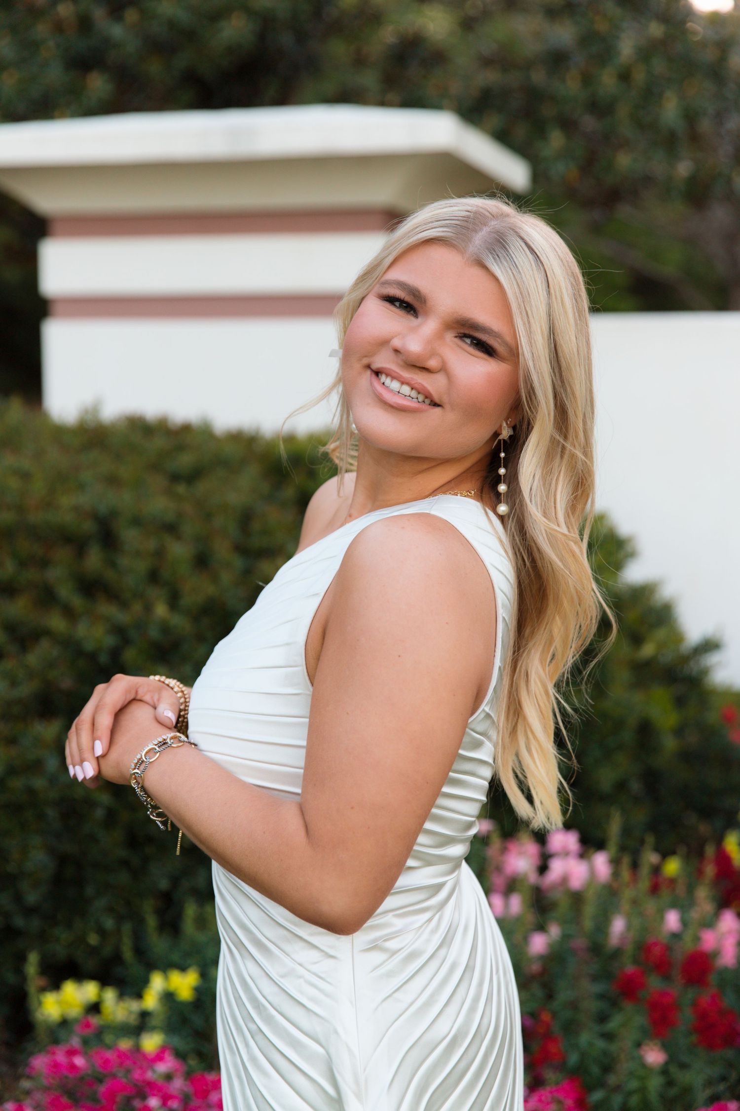 A person in a white dress poses playfully in a garden setting with pink flowers in the background.