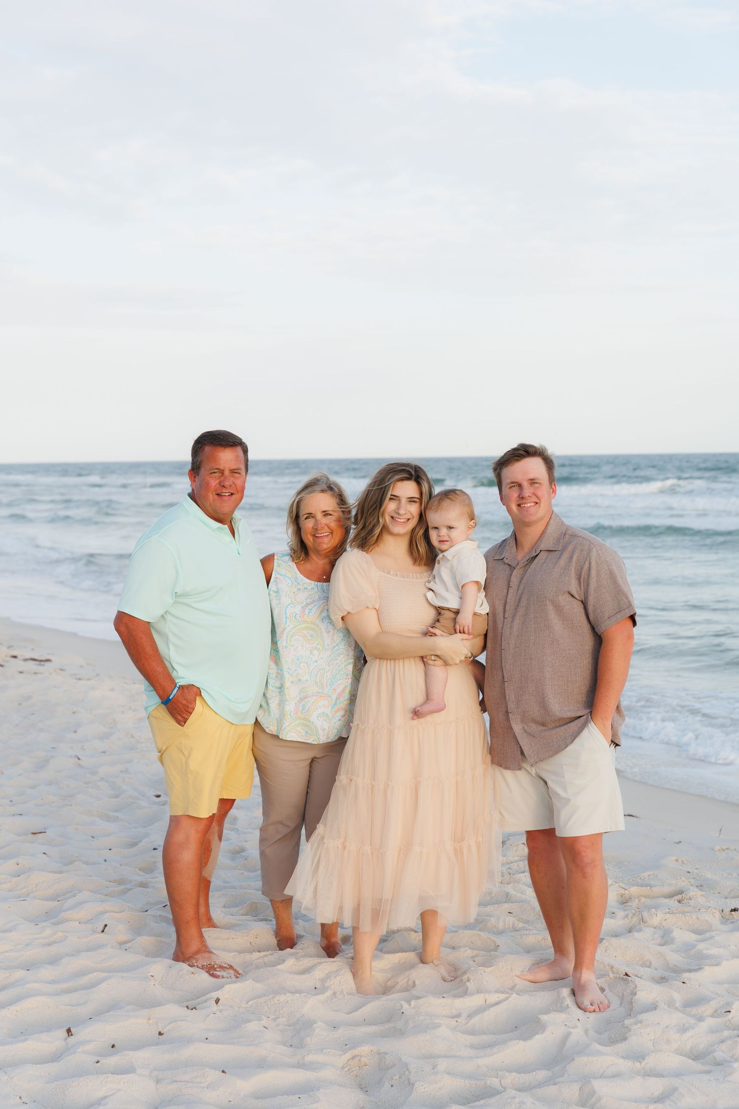 Extended family of five posing together on white sandy beach with ocean backdrop.