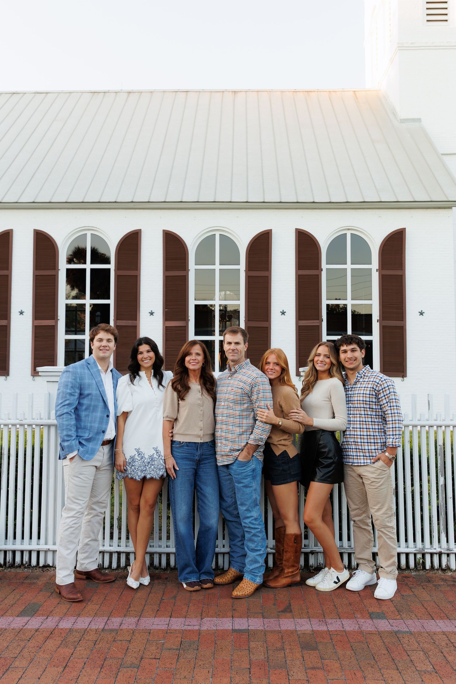 A family of seven poses together smiling in front of a white church with brown shutters on a brick pathway.