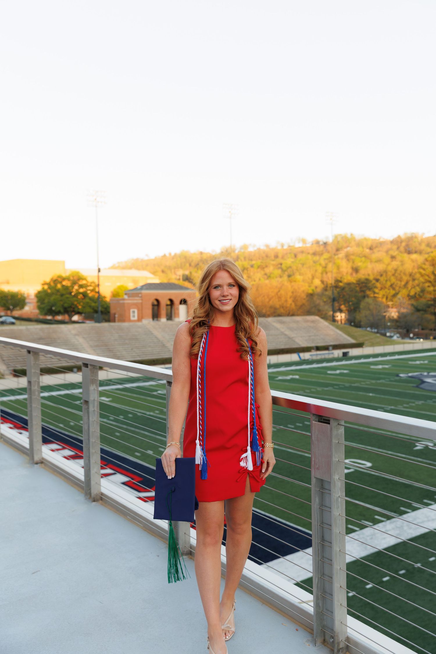 Young woman in red dress holding graduation cap with honor cords on stadium balcony.