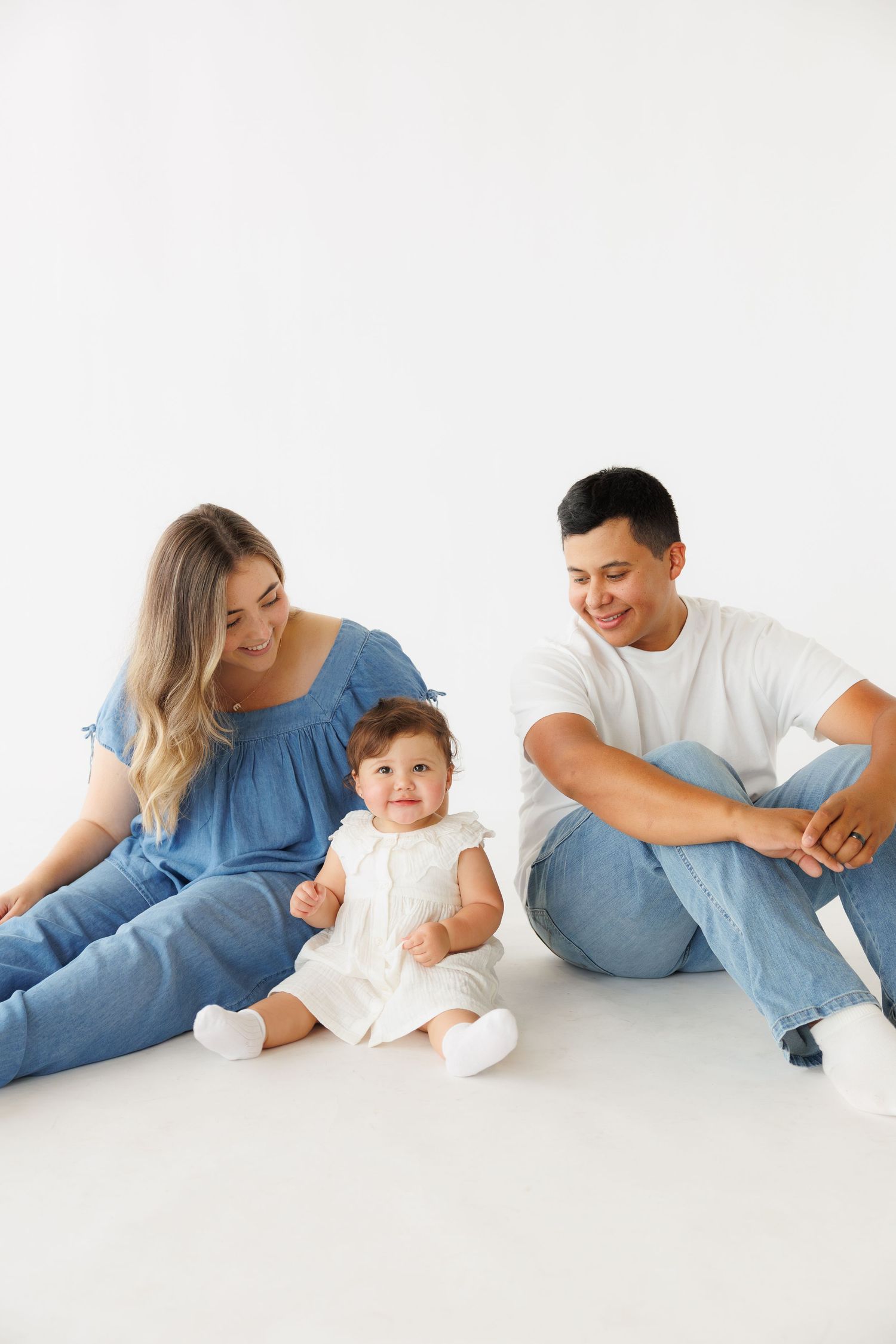 Happy young family with baby girl sitting on white floor, parents smiling at toddler in white dress.