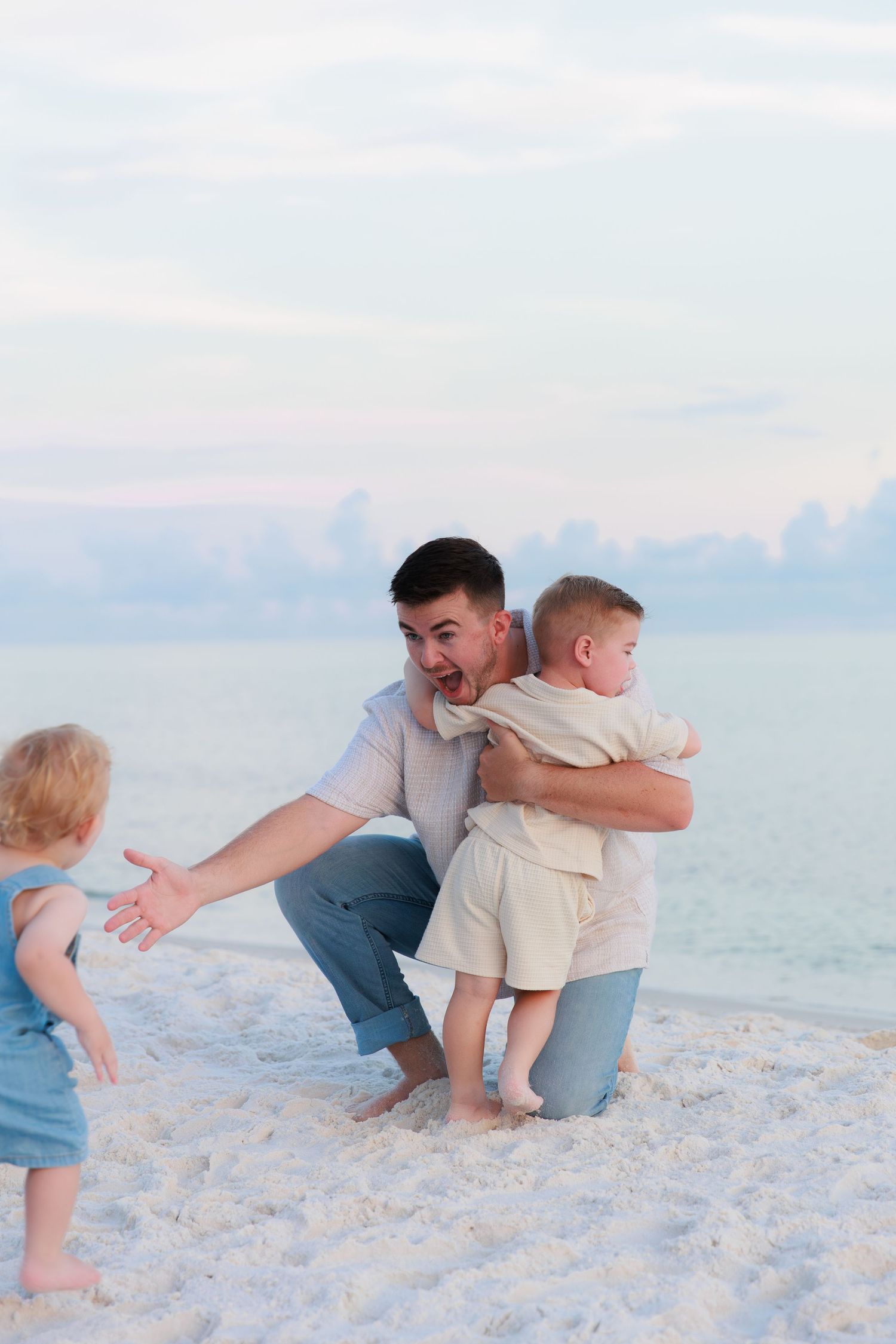 Father joyfully hugging young son on white sand beach while toddler approaches with arms out.