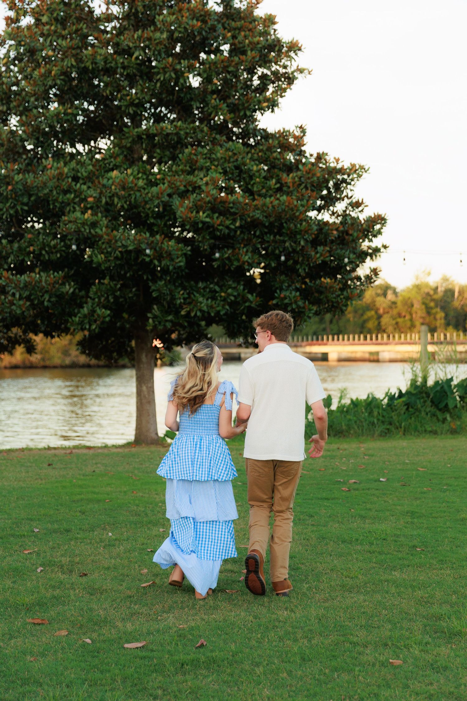 Couple walking hand in hand on green grass toward a serene waterfront, woman in blue gingham dress, man in white shirt.