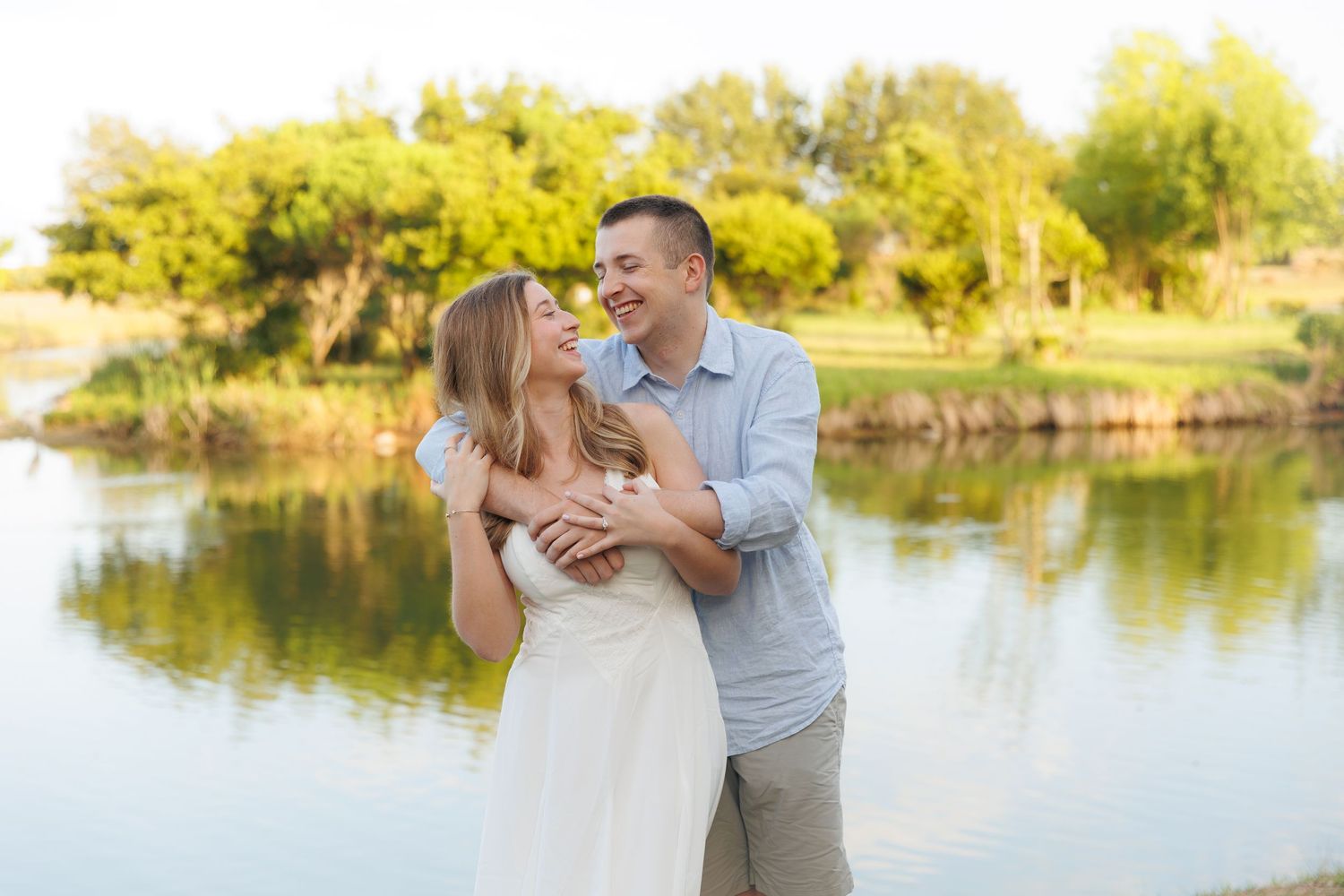 Happy couple embracing and laughing together by a serene lake surrounded by lush green trees.